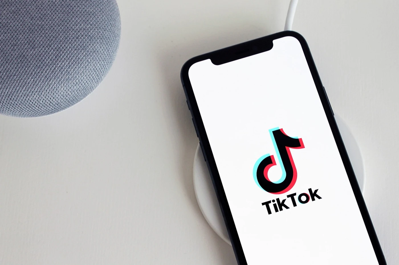 Smartphone on a wireless charger displaying the TikTok logo, next to a gray fabric smart speaker on a white surface.