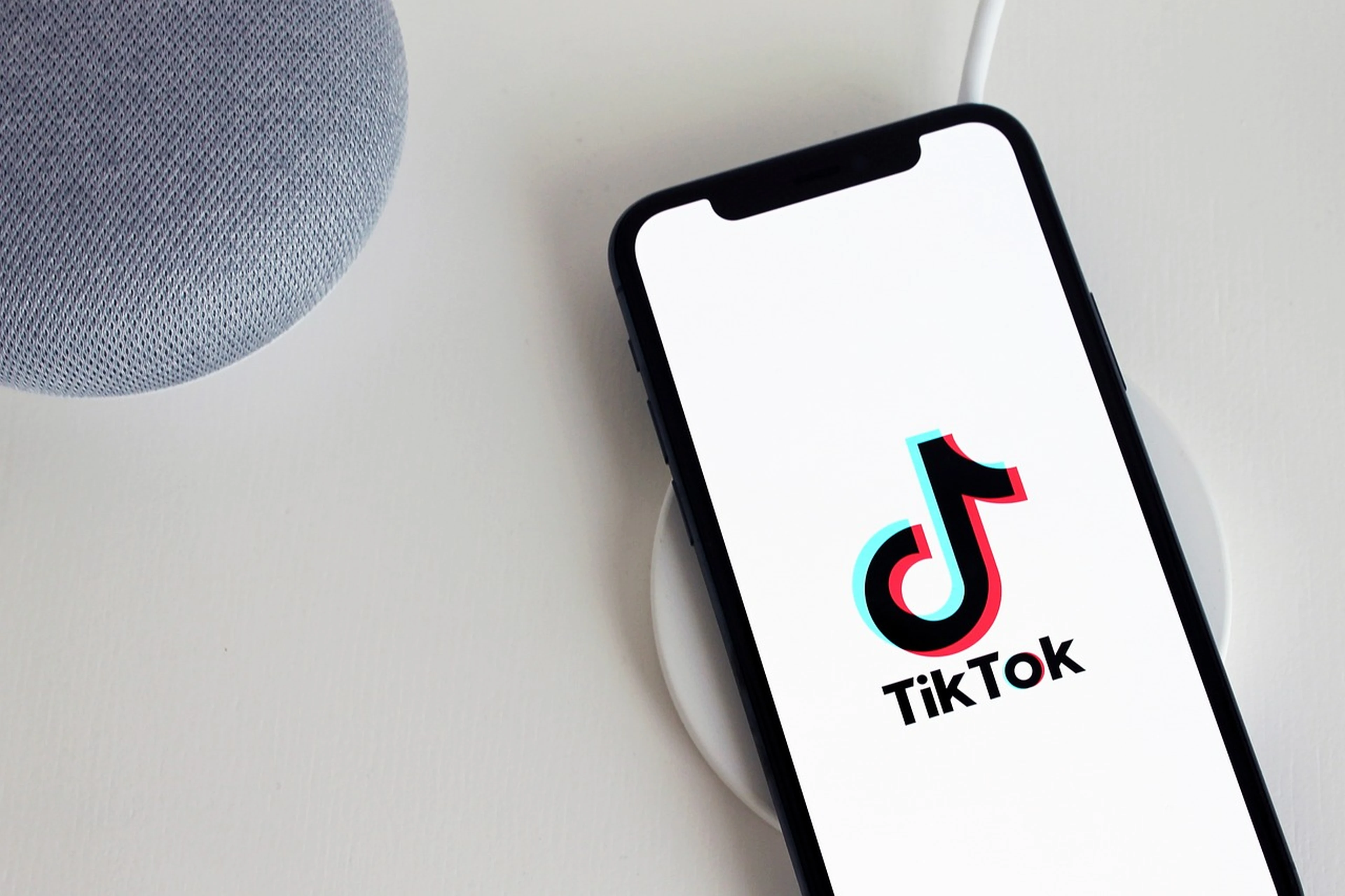 Smartphone on a wireless charger displaying the TikTok logo, next to a gray fabric smart speaker on a white surface.