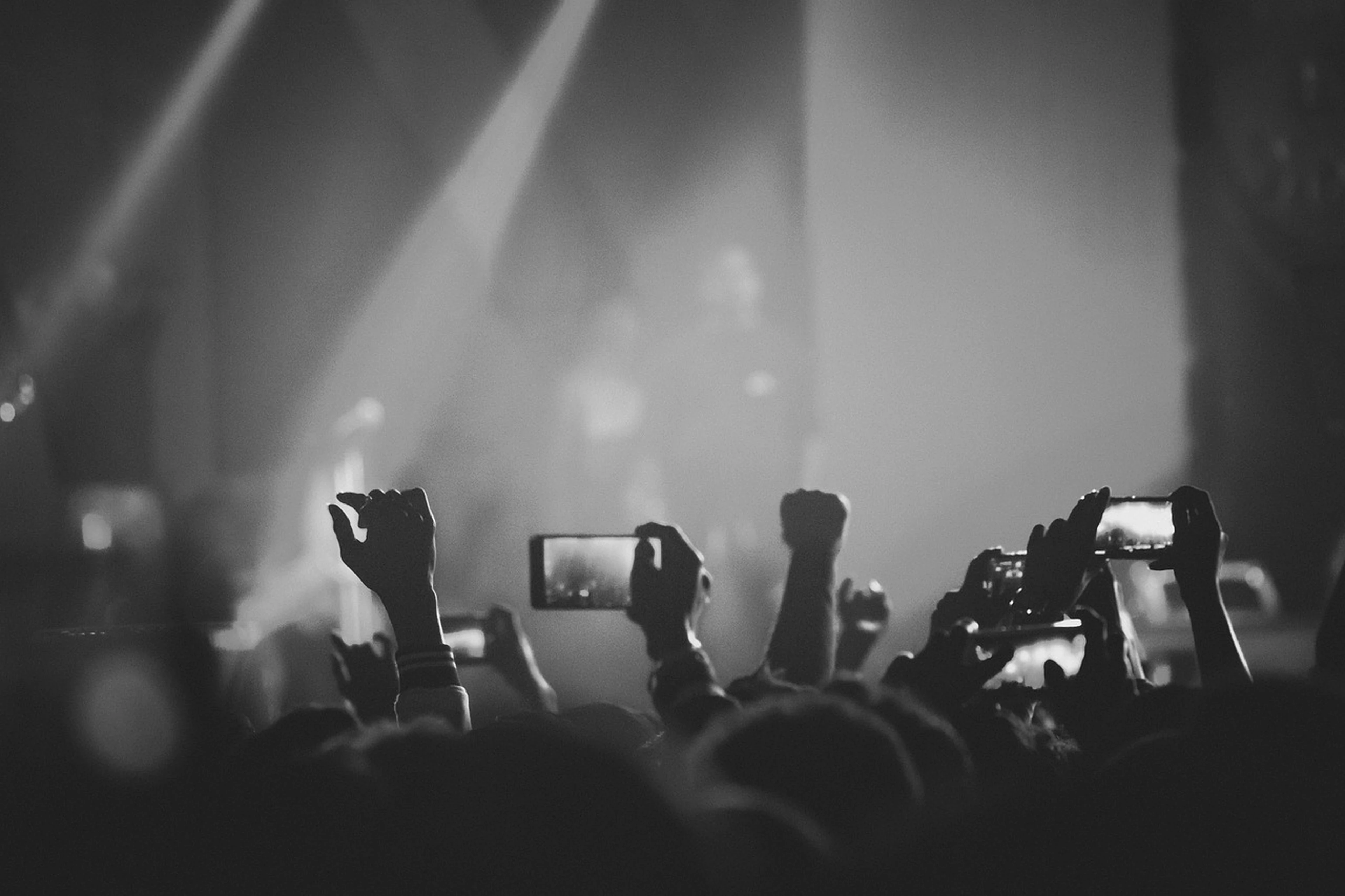 Audience at a concert, hands raised, some holding phones, capturing the moment. Stage lights create a dramatic atmosphere in black and white.