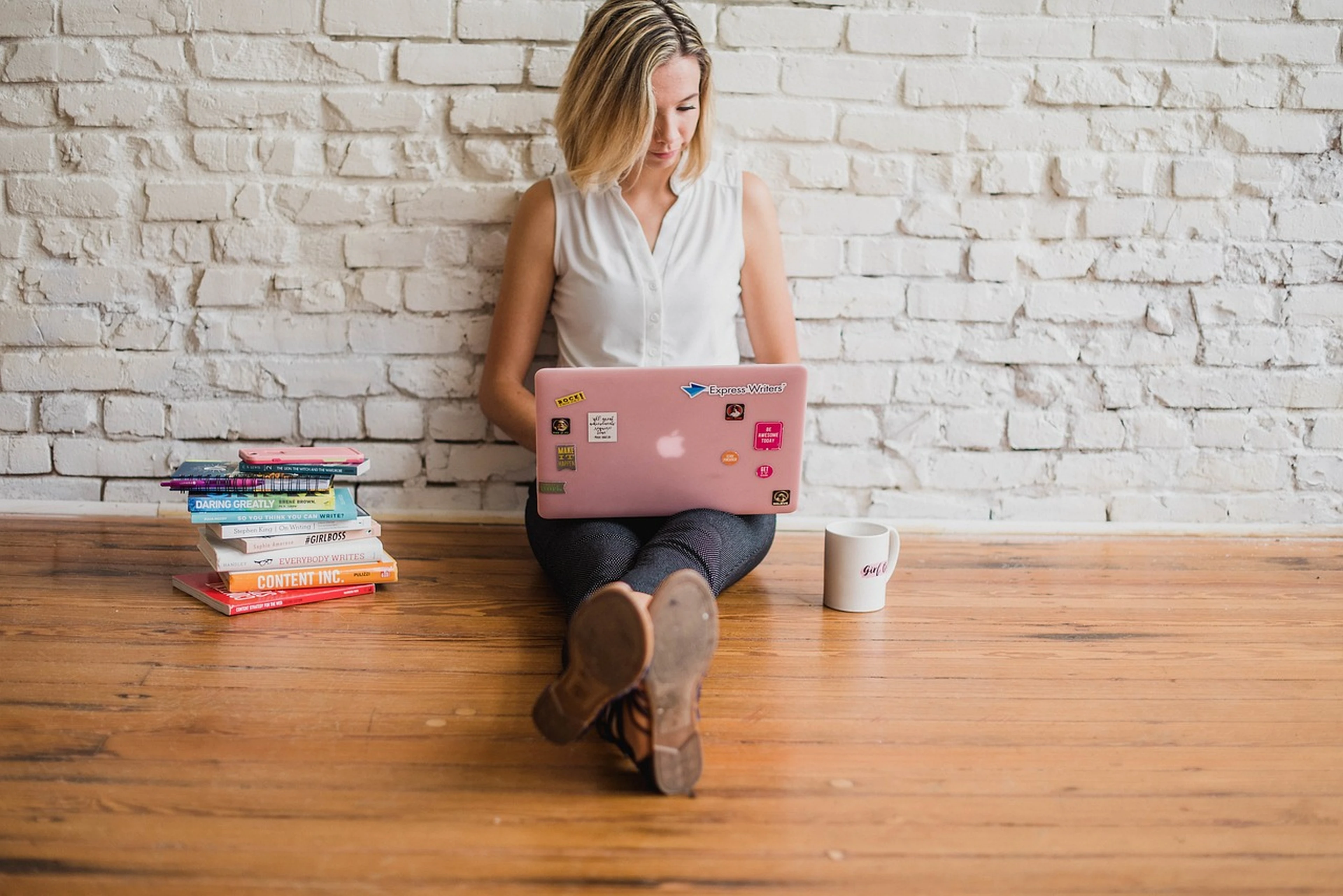 A woman influencer sitting on a wooden floor, using a pink laptop to upload pictures from her solo travel