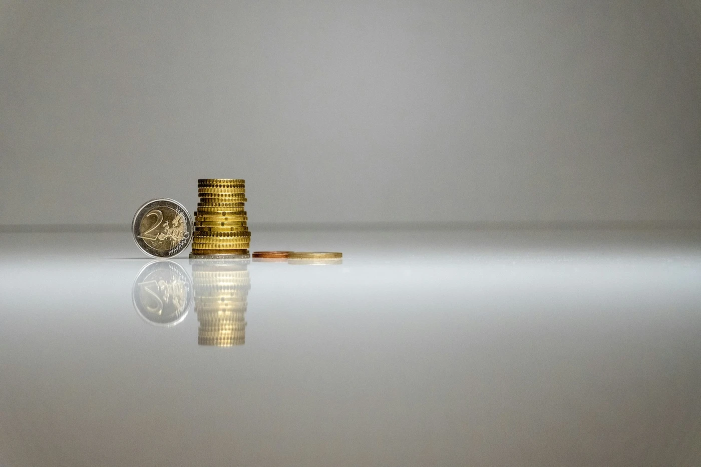 Coins stacked and scattered on a reflective surface, including a visible two-euro coin, against a plain gray background.