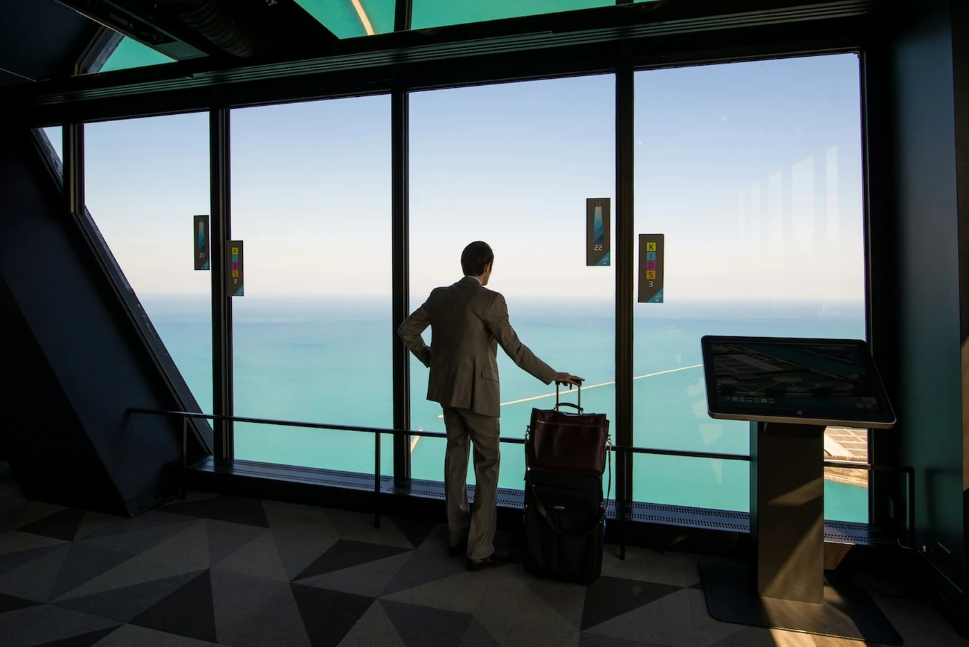 A person in a suit stands with luggage, looking out from a large window at a vast ocean view under a clear sky.
