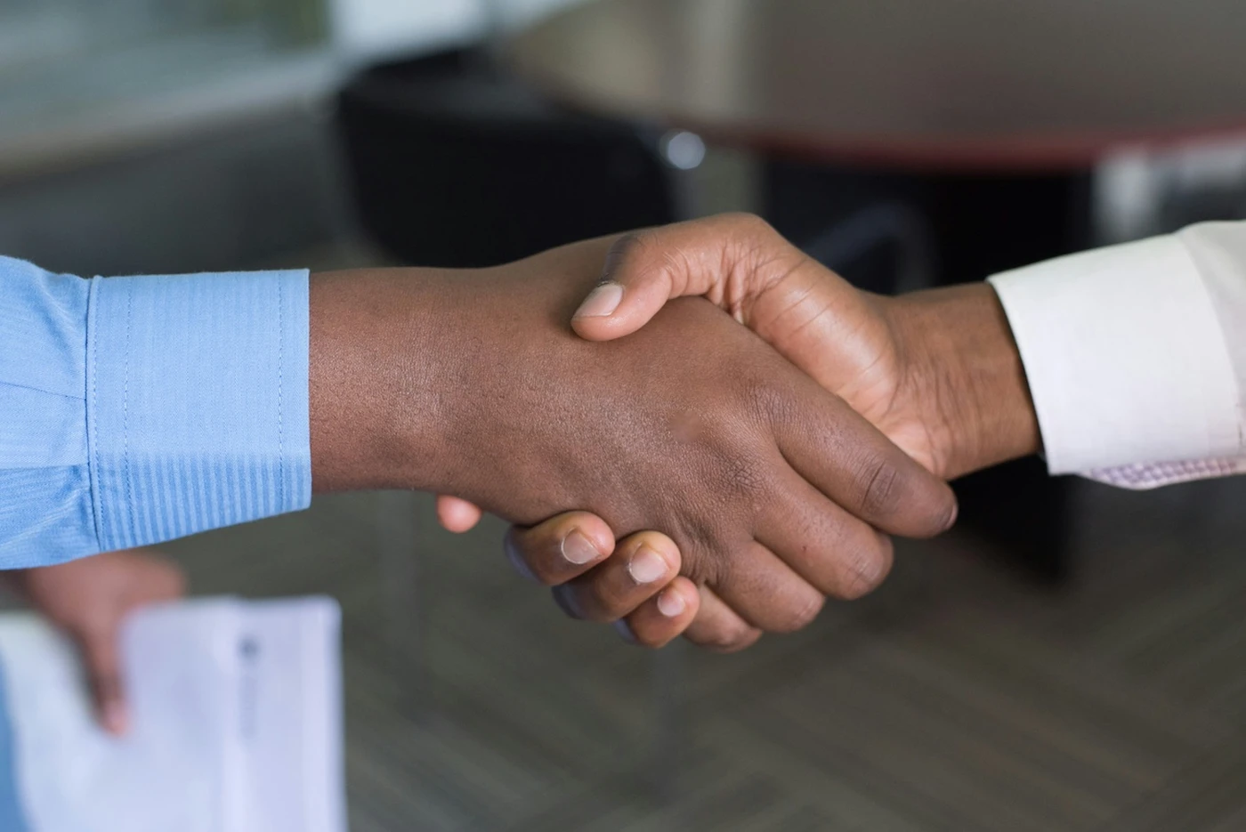 Two people shaking hands, one wearing a blue shirt and the other a white shirt, in an office setting.