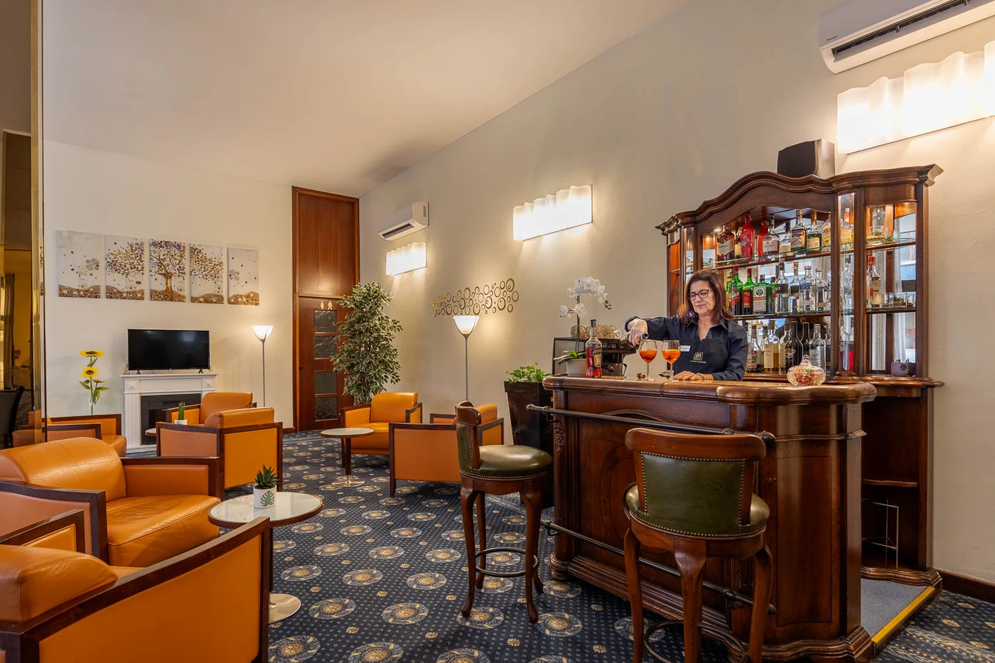 Cozy hotel lounge with wooden bar, bartender serving cocktails, orange armchairs, blue patterned carpet, wall lamps, TV and plants.