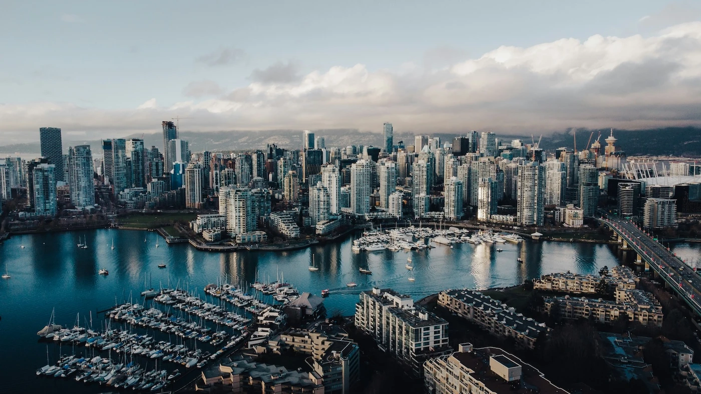 Aerial view of Vancouver skyline with glass towers, marinas full of boats, calm blue water, and distant mountains under a cloudy sky.