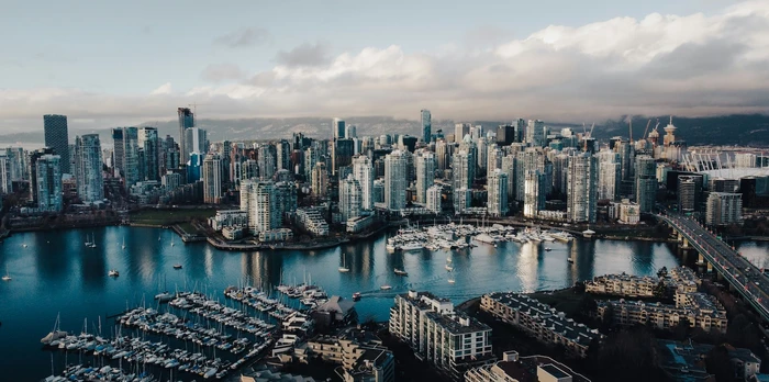 Aerial view of Vancouver skyline with glass towers, marinas full of boats, calm blue water, and distant mountains under a cloudy sky.