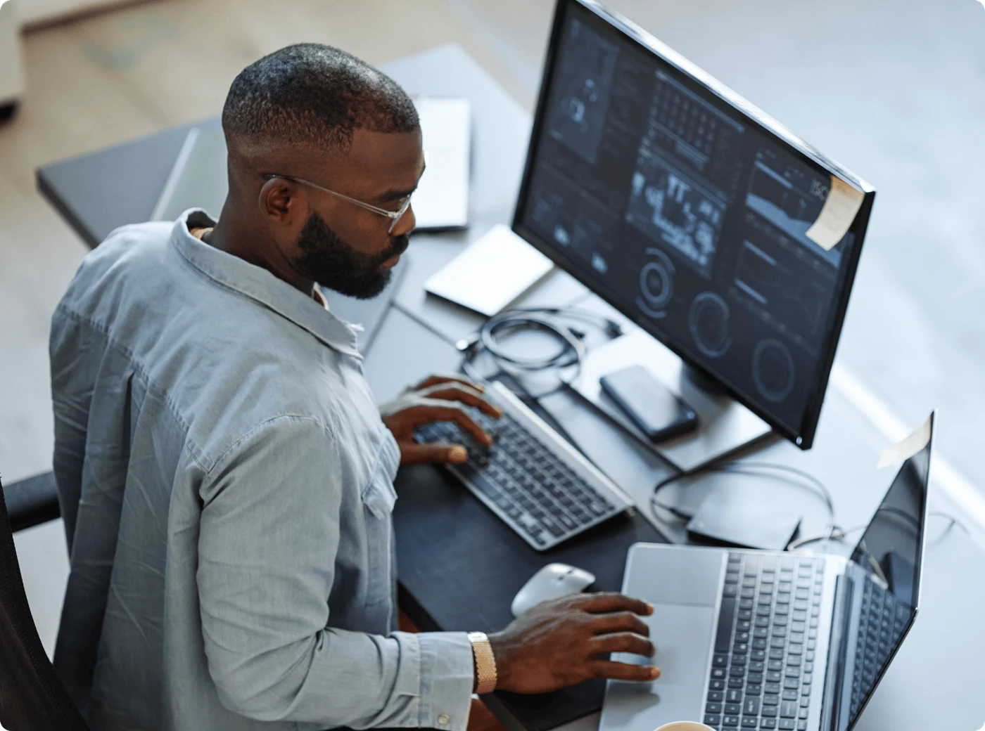 Person at a desk uses a laptop and external monitor showing charts and dashboards, typing on a keyboard with mouse and cables nearby.