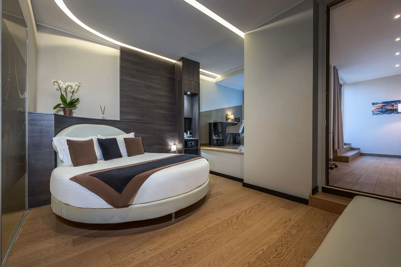 Contemporary hotel room with round bed, wood paneling and floors, cove lighting, wall mirror, TV, orchids, and a raised adjoining area.