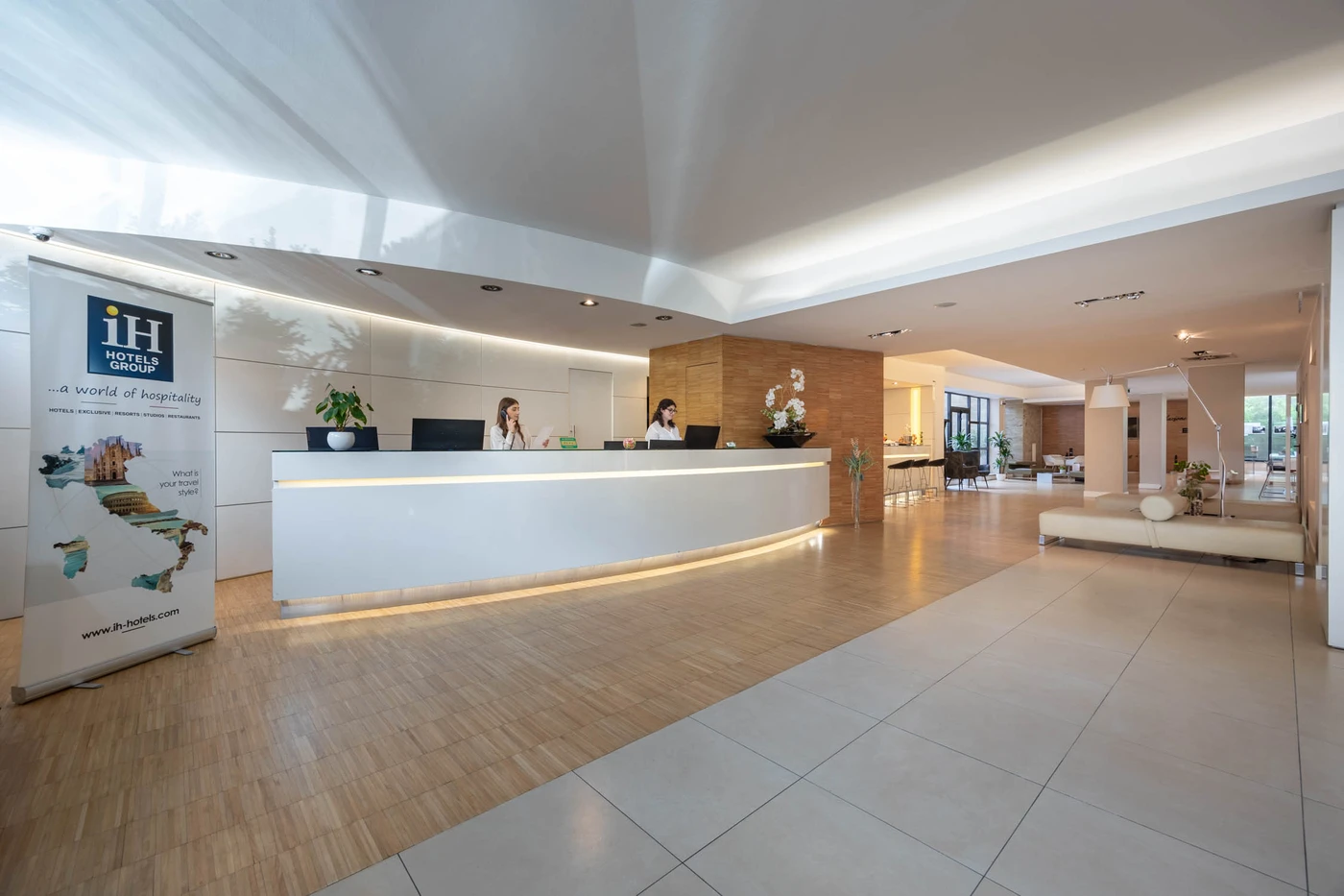 Modern hotel lobby with curved white reception desk, two staff, wood and tile floors, plants, seating area, and an IH Hotels banner.