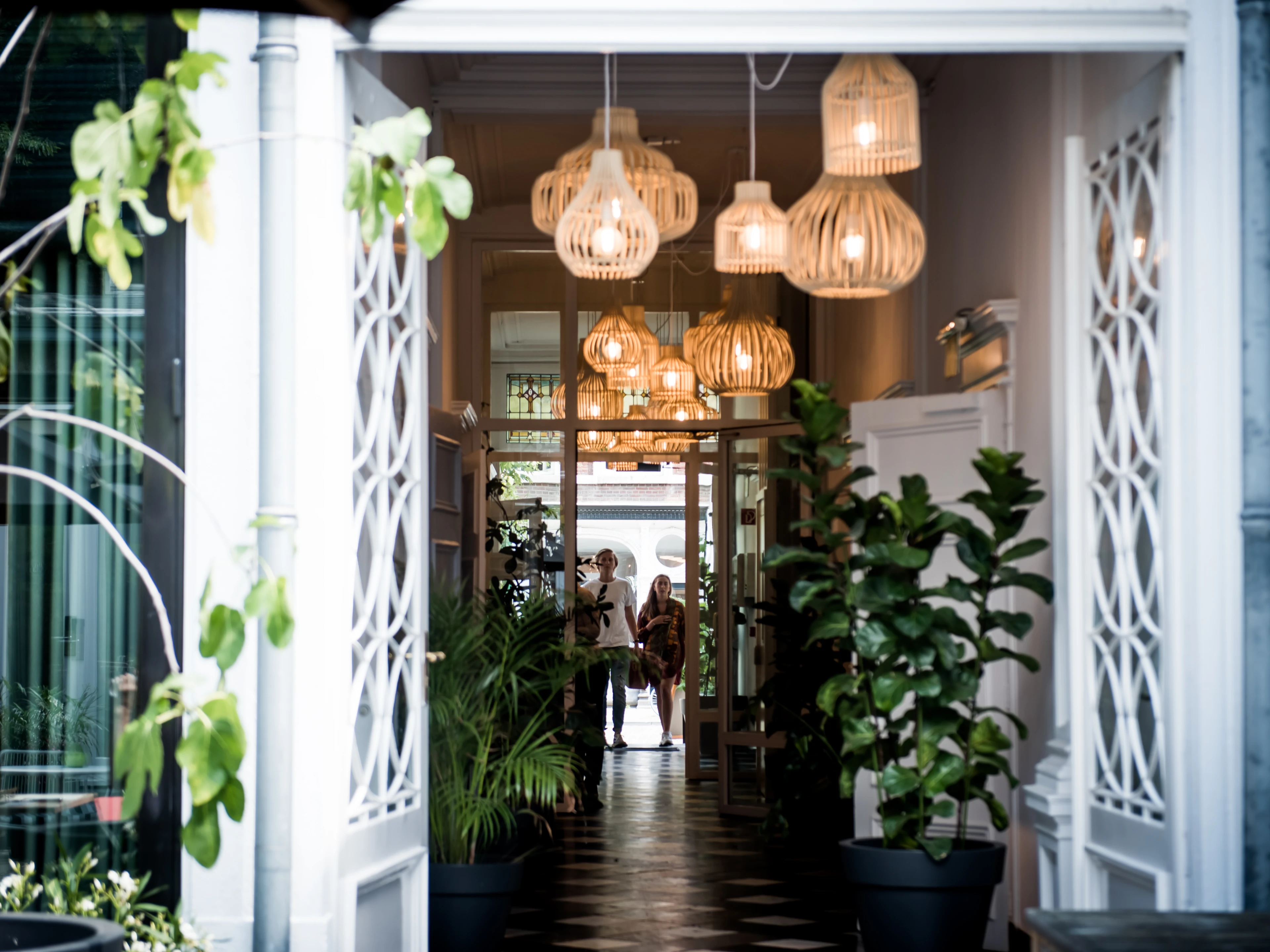 Open doorway into a stylish interior with hanging rattan lamps, lush potted plants and two people standing in the back.