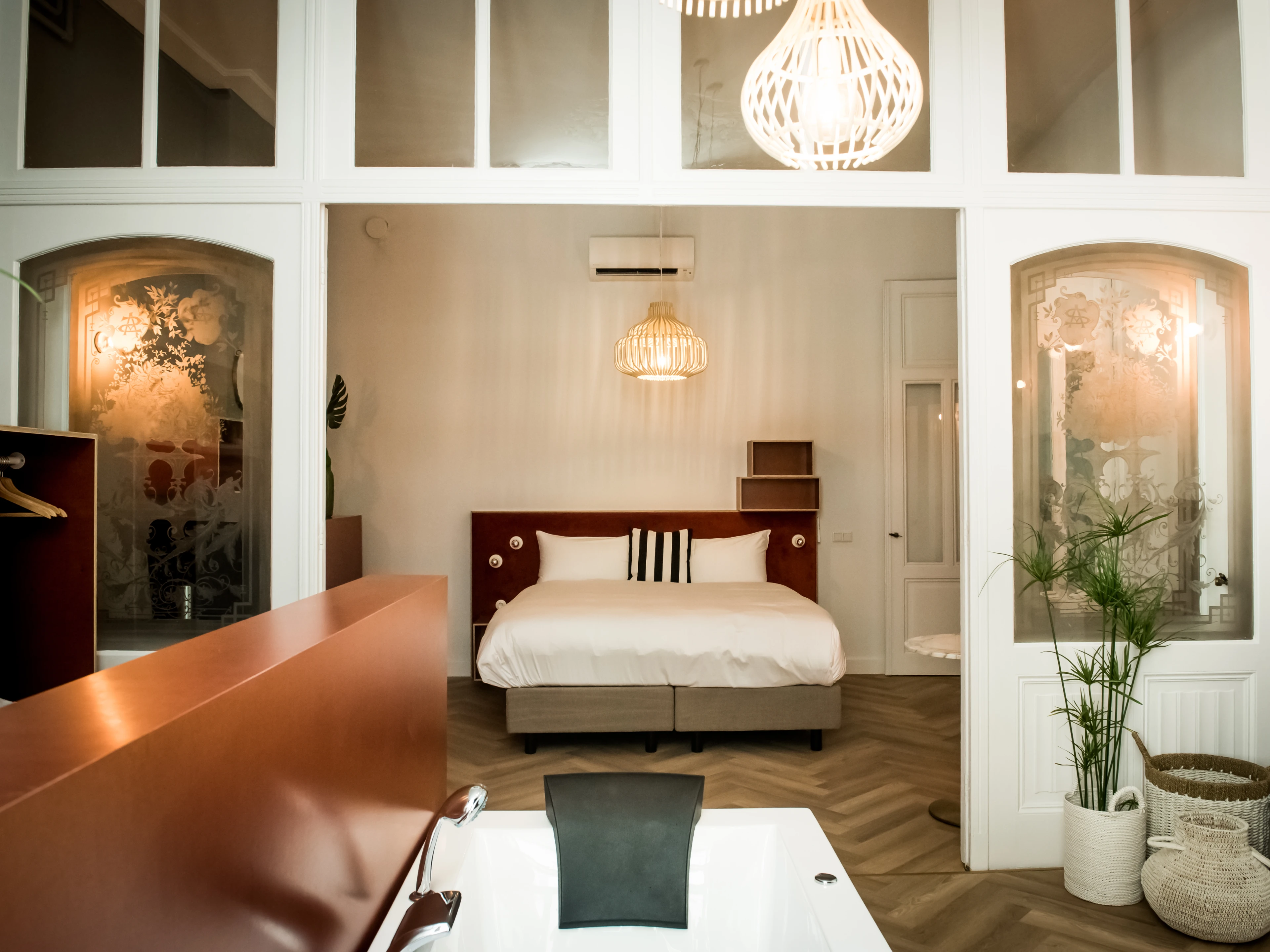 Modern bedroom with white bed, pendant lights, glass partitions, potted plants and a freestanding bathtub in the foreground.