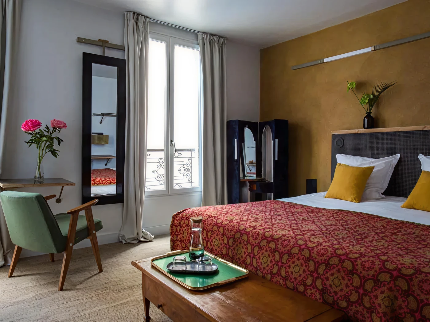 Bright hotel bedroom with patterned red bedspread, yellow pillows, wooden bench with tray, window with curtains, mirror and vase of pink roses.
