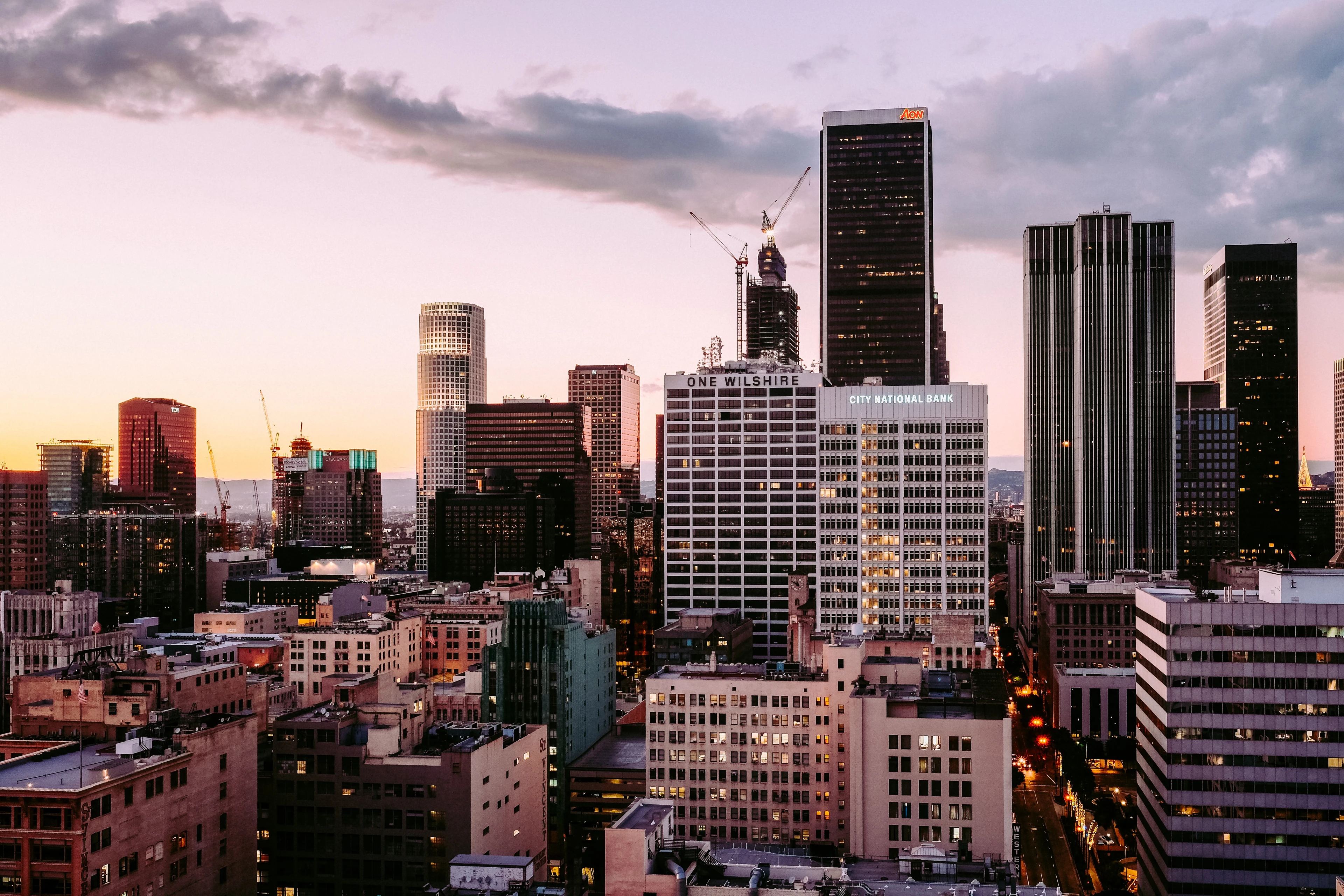 Aerial view of a city skyline at dusk, with tall buildings, cranes, and a pinkish sky. The streets are illuminated with lights.