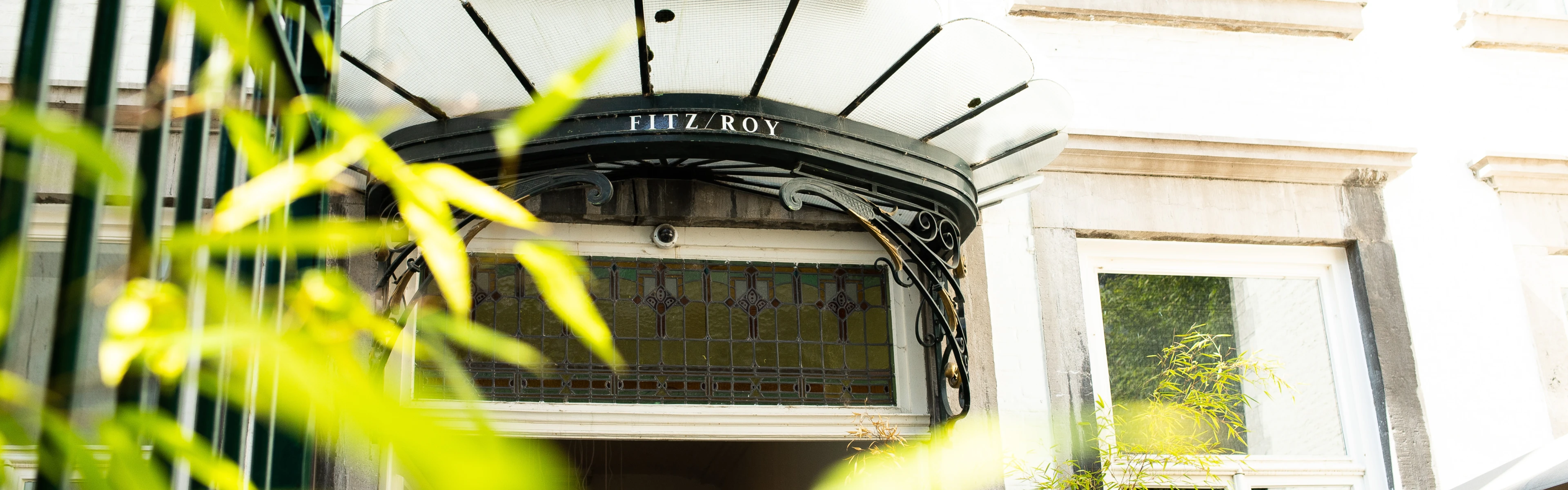 Ornate iron-and-glass canopy reading "Fitz Roy" over a pale building entrance with stained-glass transom and blurred foliage in the foreground.