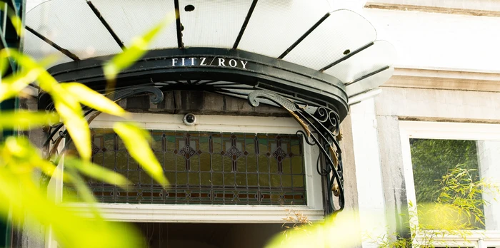 Ornate iron-and-glass canopy reading "Fitz Roy" over a pale building entrance with stained-glass transom and blurred foliage in the foreground.