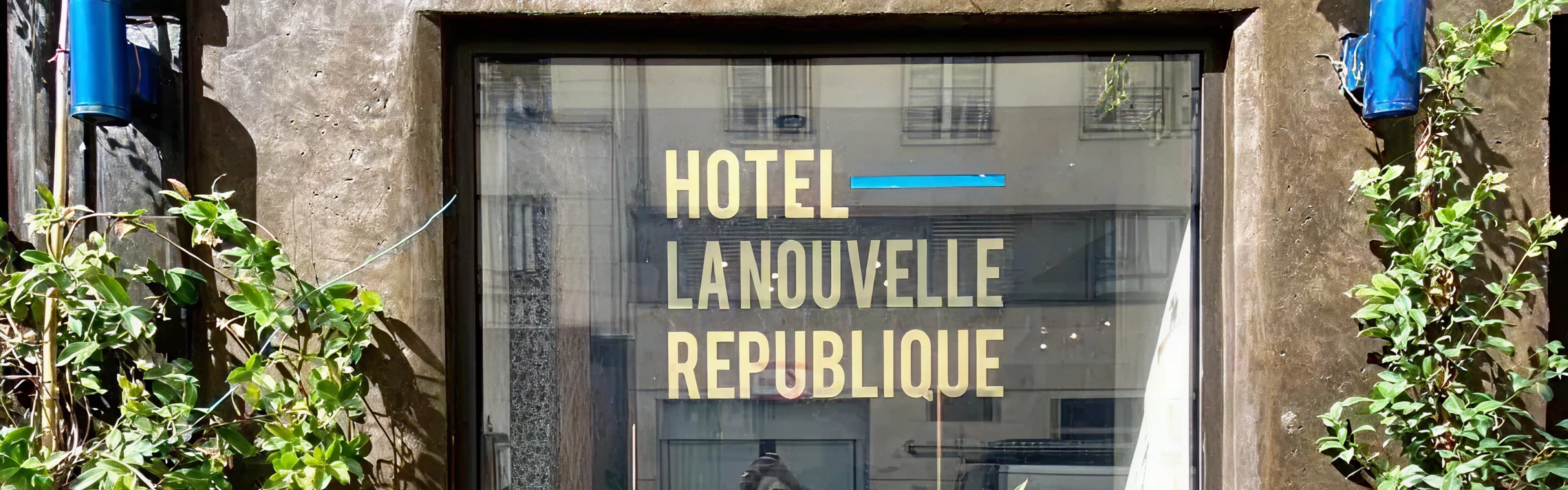 Glass hotel entrance window with gold lettering 'HOTEL LA NOUVELLE RÉPUBLIQUE', flanked by green climbing plants on a weathered stone facade.