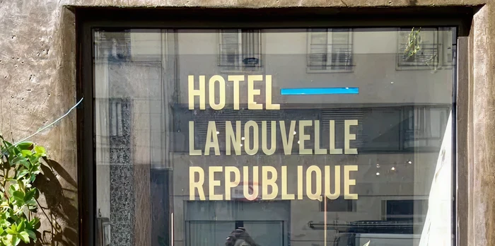 Glass hotel entrance window with gold lettering 'HOTEL LA NOUVELLE RÉPUBLIQUE', flanked by green climbing plants on a weathered stone facade.