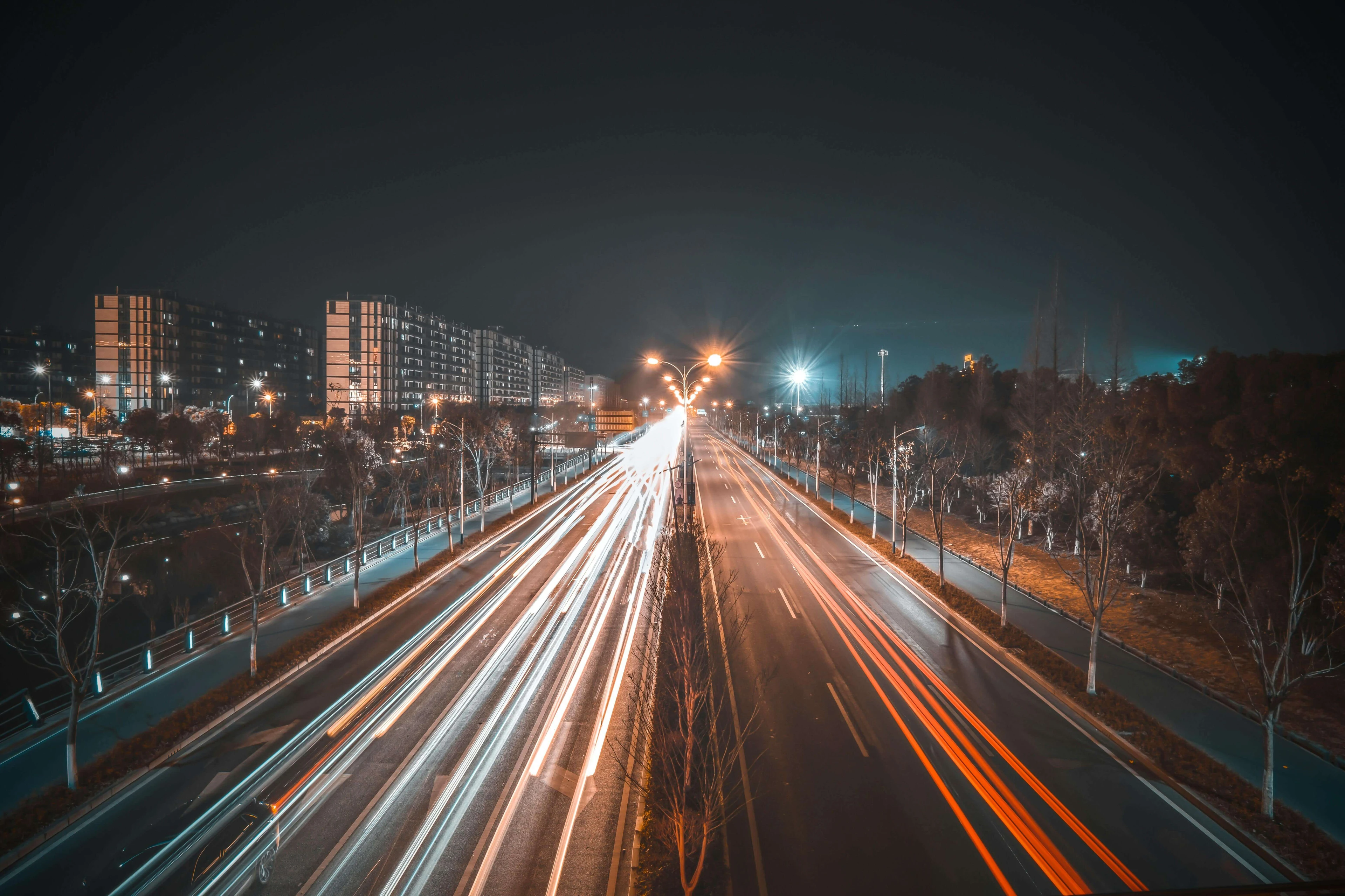 Long-exposure night highway with bright white and red light trails, apartments on the left and trees along the right side.