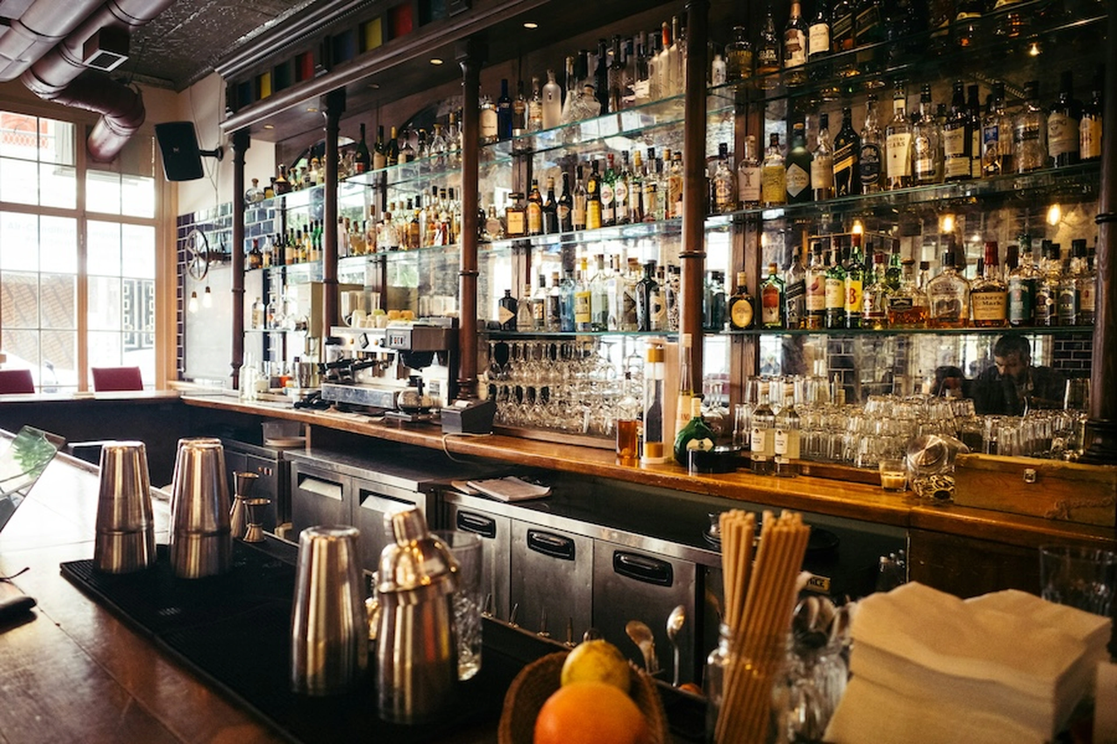 Well-stocked bar with mirrored shelves of spirits, wooden counter with shakers, fruit, and glassware, warm daylight through tall windows.