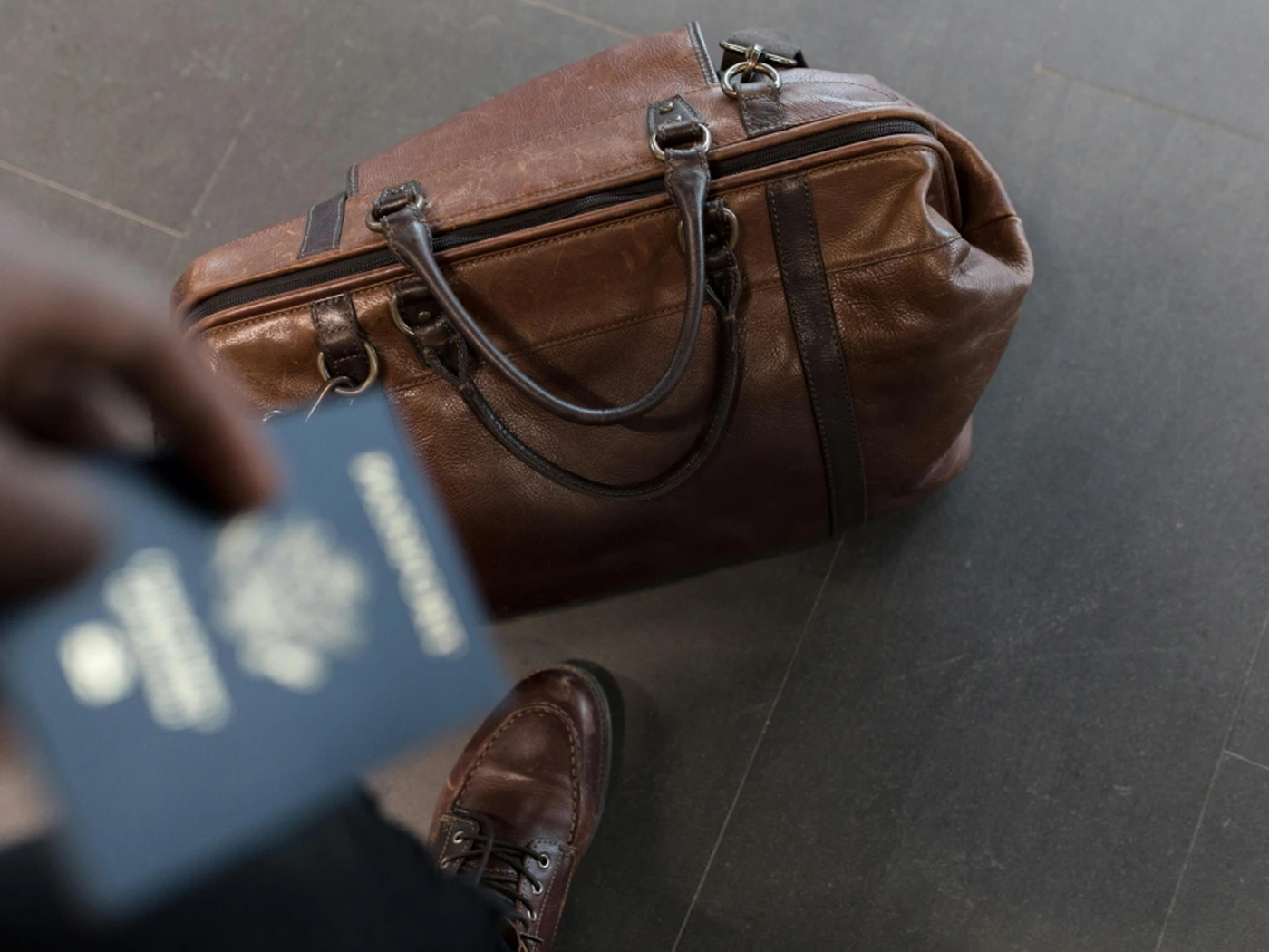 A brown leather duffel bag on the floor next to a person holding a blue passport, wearing brown leather shoes.