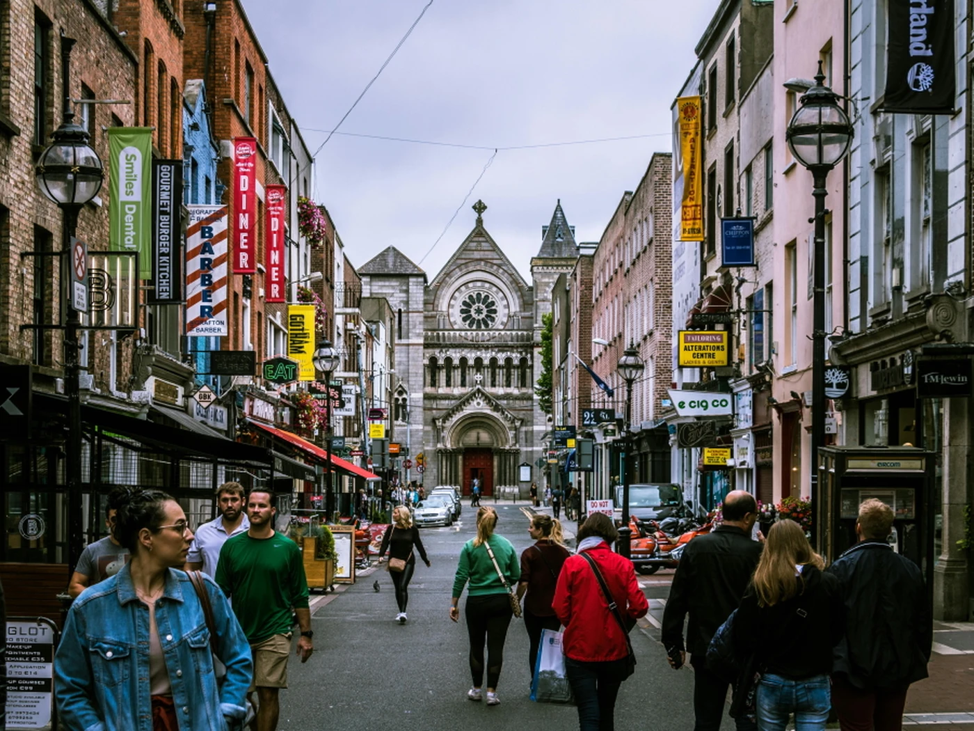 A bustling street with people walking, lined with colorful shops and signs, leading to a historic stone church with a large rose window.