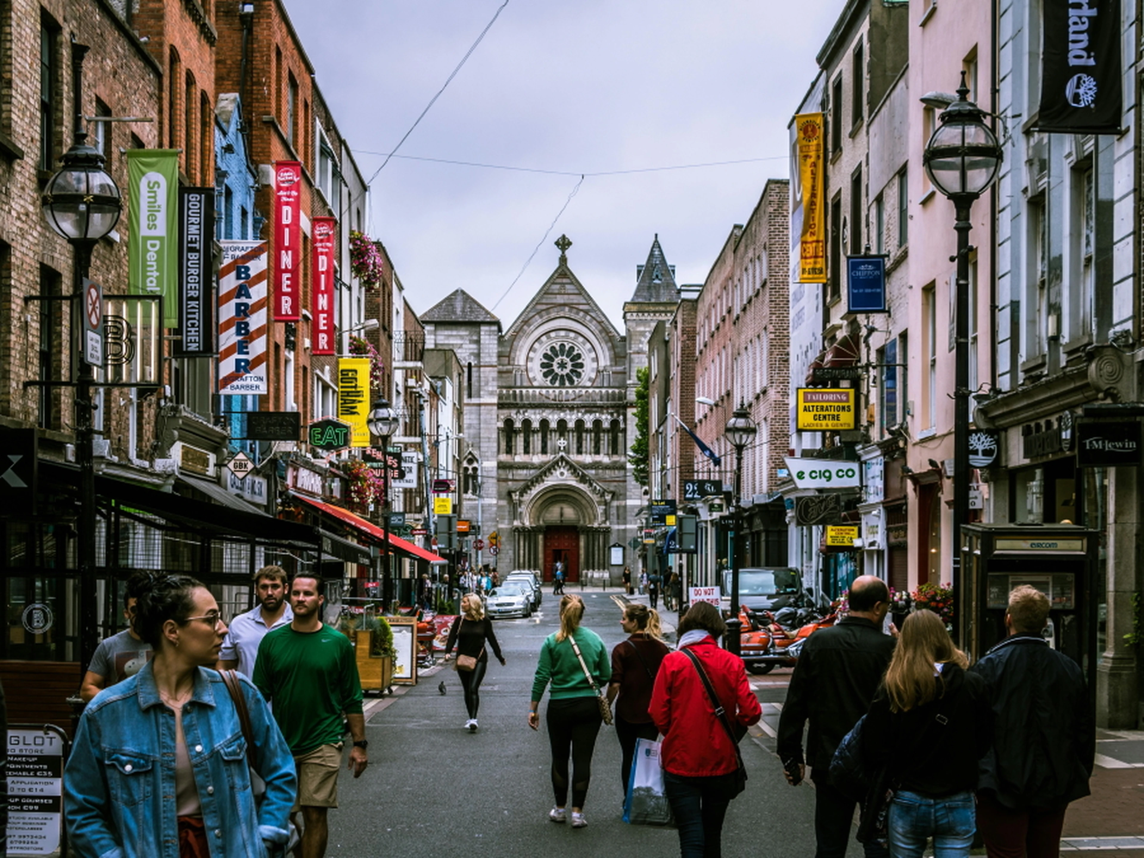 A bustling street with people walking, lined with colorful shops and signs, leading to a historic stone church with a large rose window.