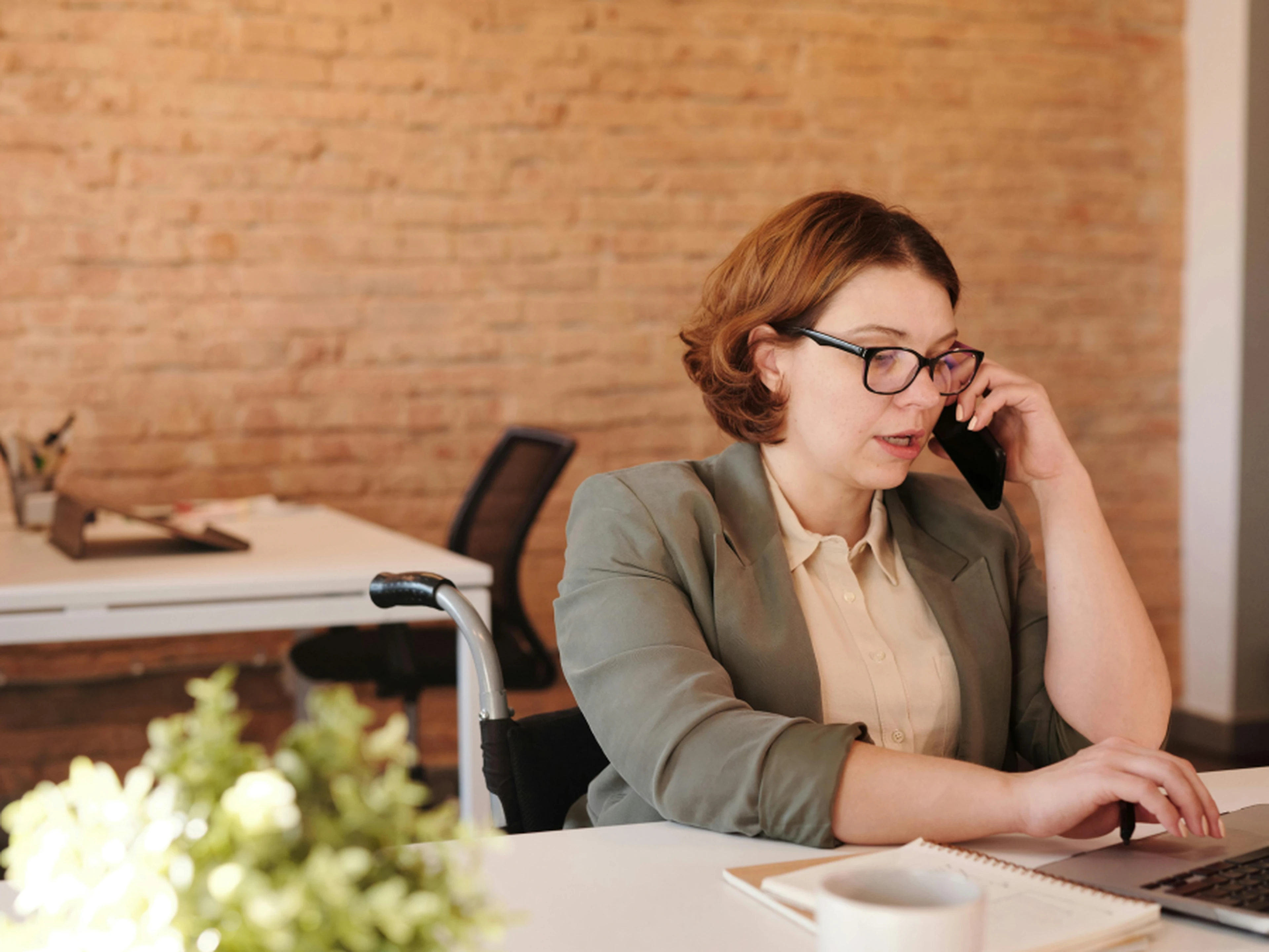 Woman. talking on a phone while typing on a laptop in an office with brick walls.