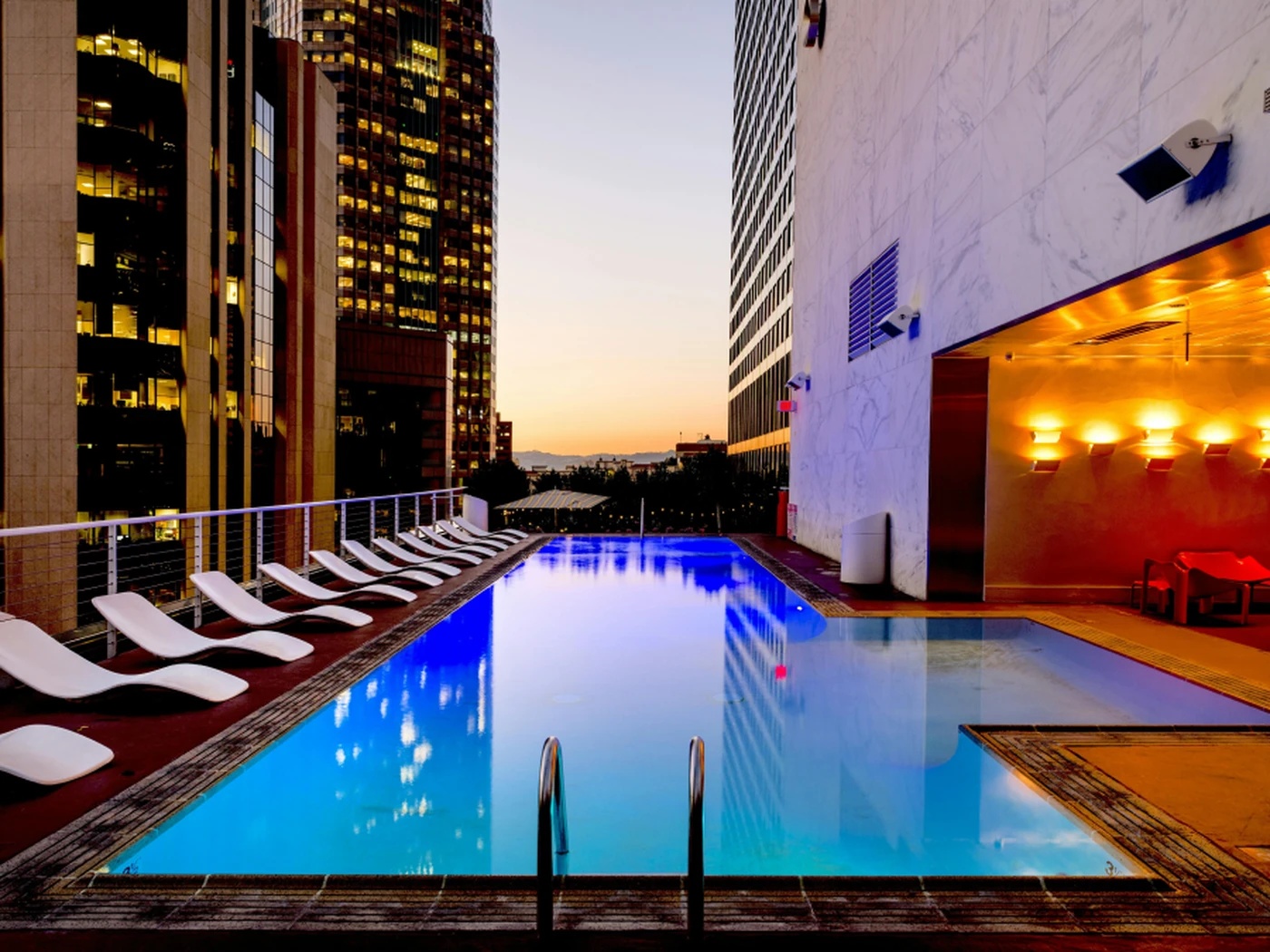Rooftop pool at sunset with modern lounge chairs, surrounded by tall buildings, and city lights in the background.