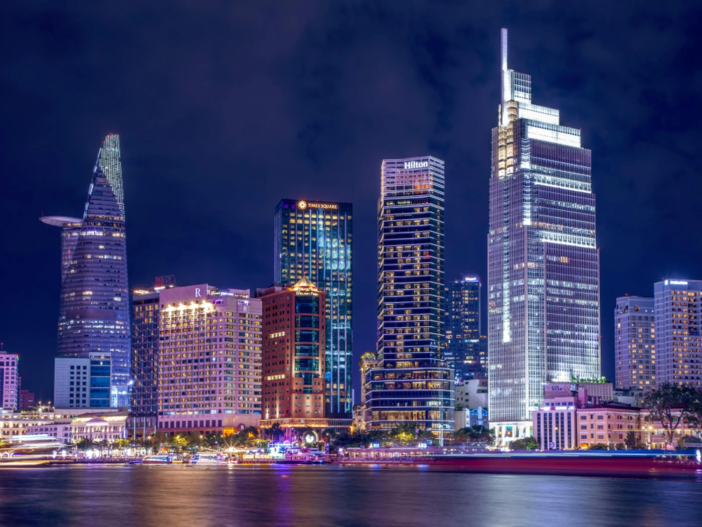 Night view of a city skyline with illuminated skyscrapers reflecting on a calm river, showcasing modern architecture against a dark sky.
