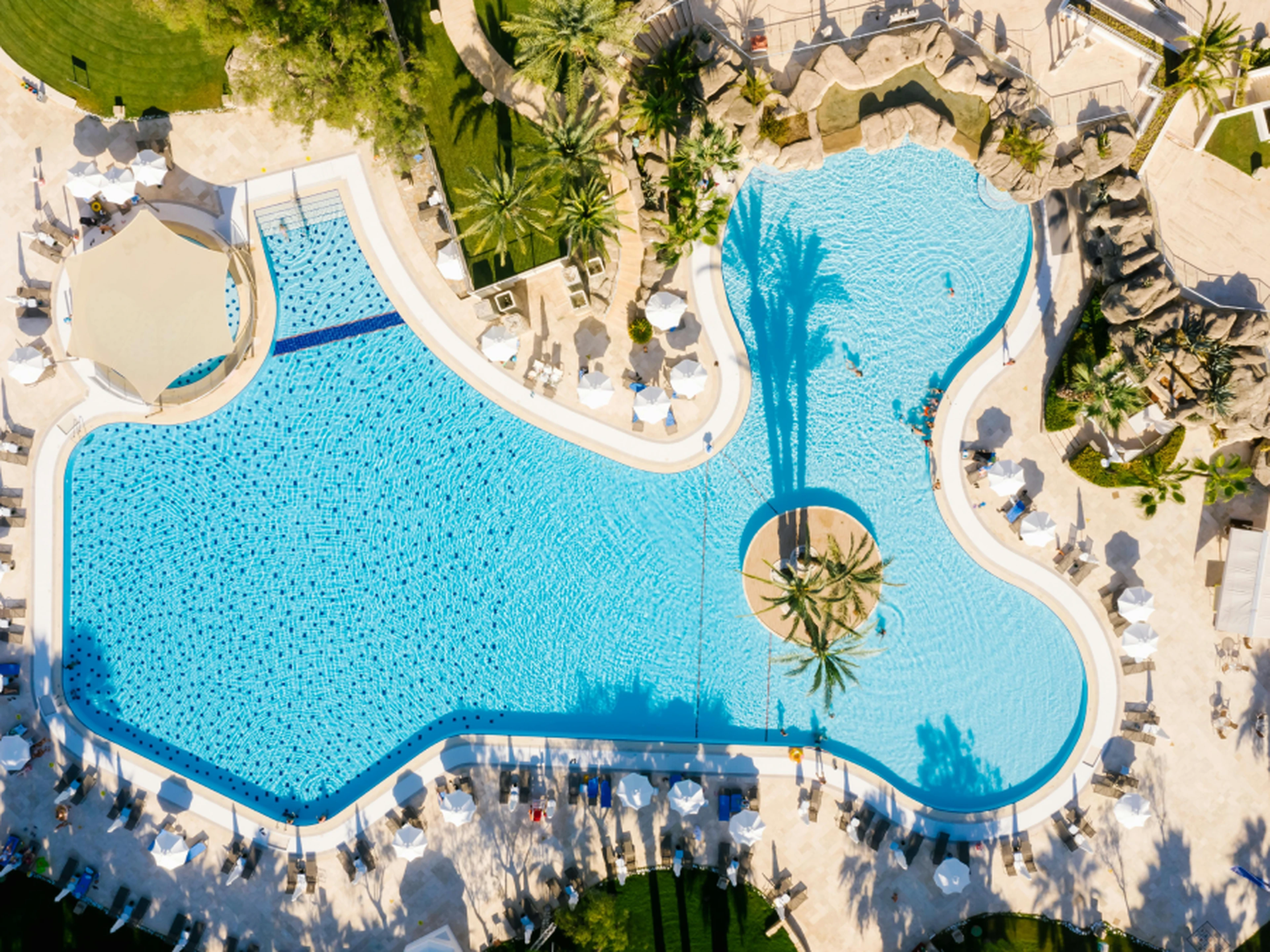 Aerial view of a large, uniquely shaped swimming pool with a central island, surrounded by lounge chairs and palm trees.