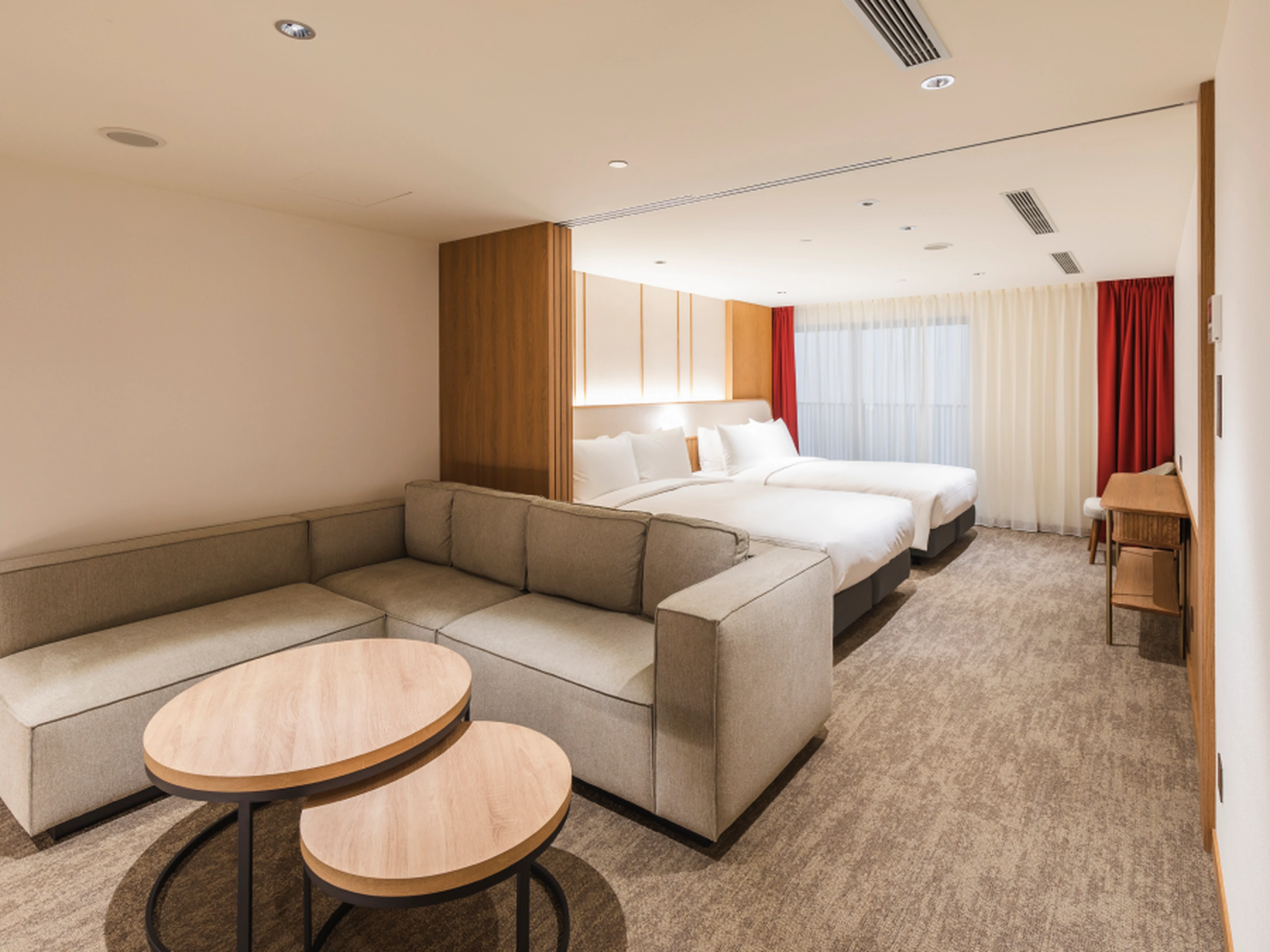 Modern hotel suite with two large beds, an L-shaped sofa, nested round coffee tables, carpeted floor, and large window with sheer and red curtains.