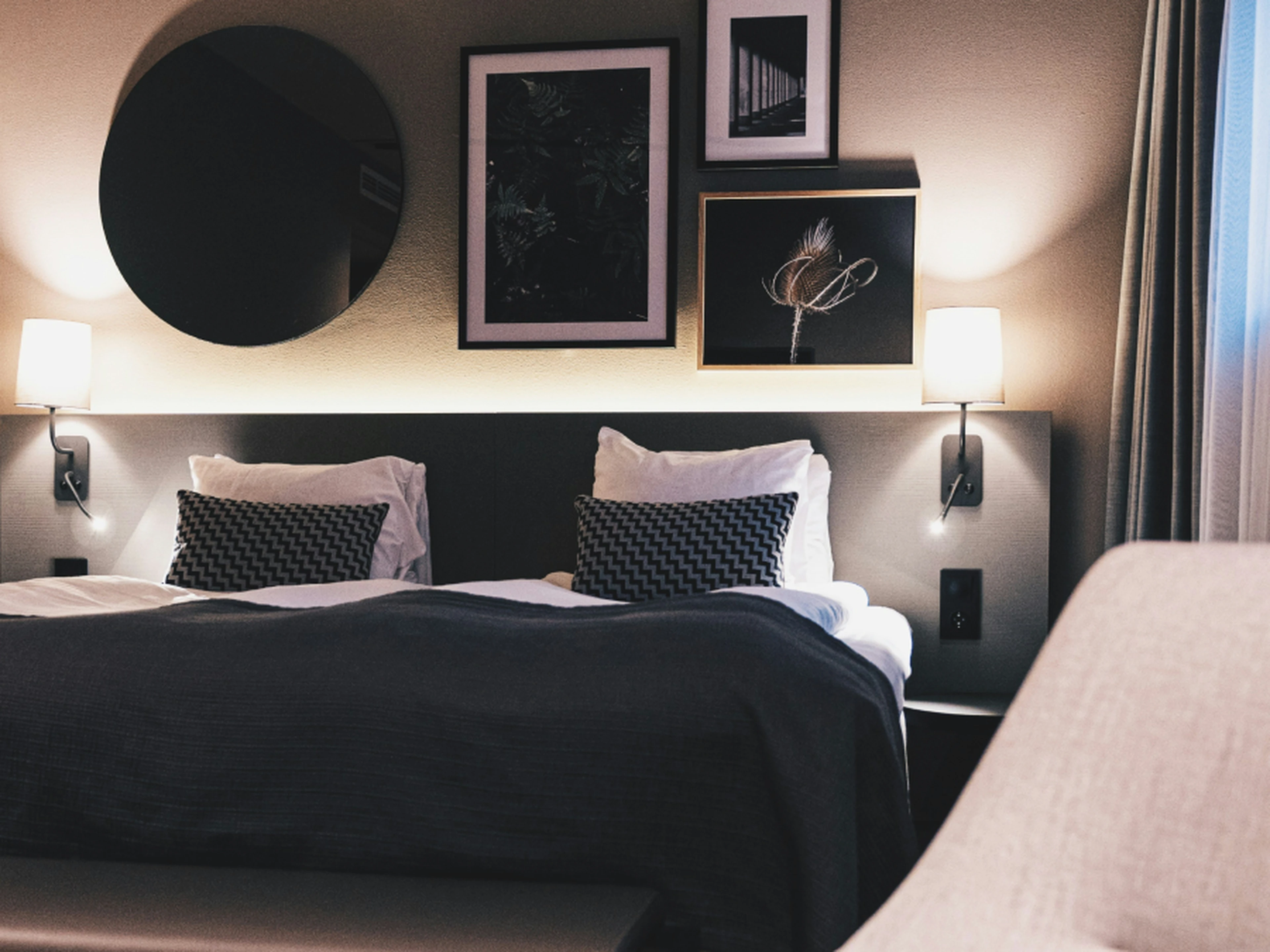Modern hotel room with two twin beds, decorative pillows, wall art, and soft lighting. A round mirror and framed pictures adorn the wall.