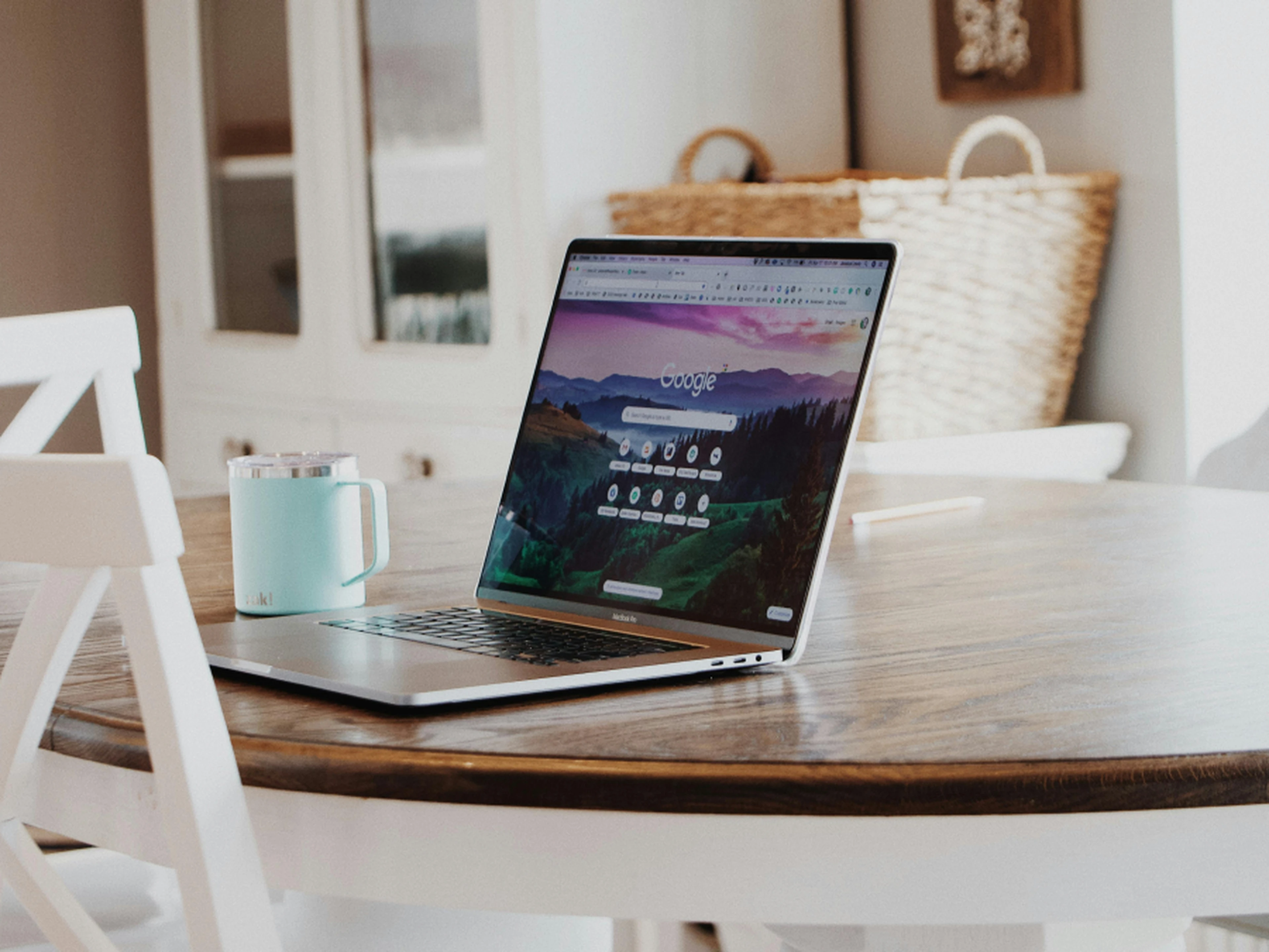 Laptop on a wooden table displaying a search engine homepage, with a blue mug nearby in a bright, cozy room.