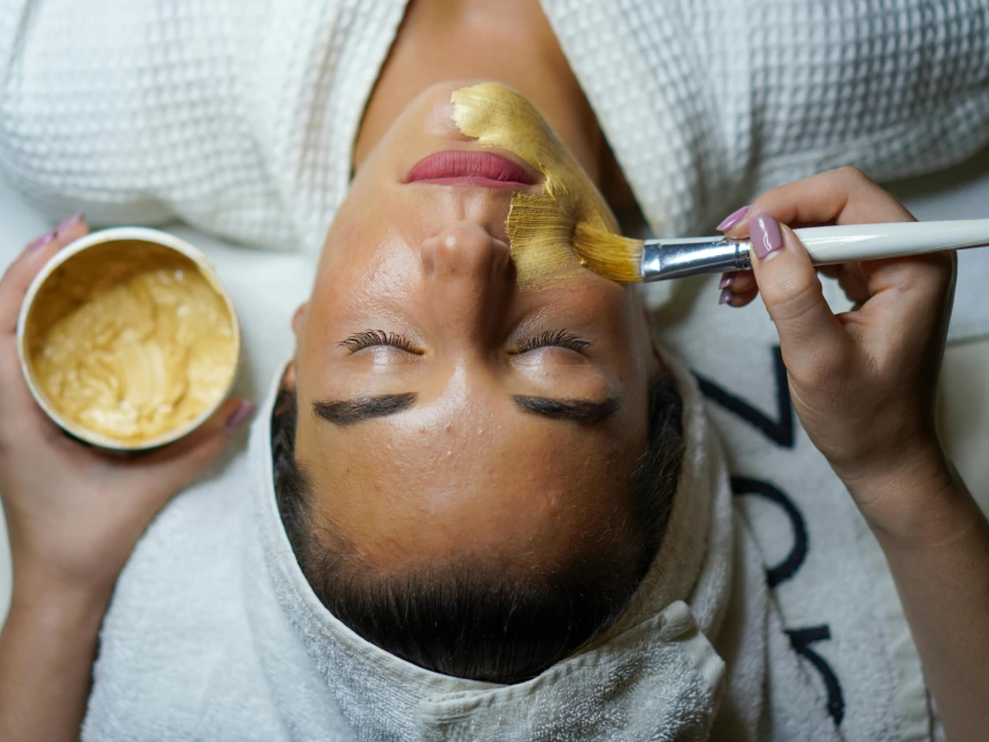 Person receiving a facial treatment, with a brush applying a golden mask. They are lying down in a spa setting, wearing a white robe.