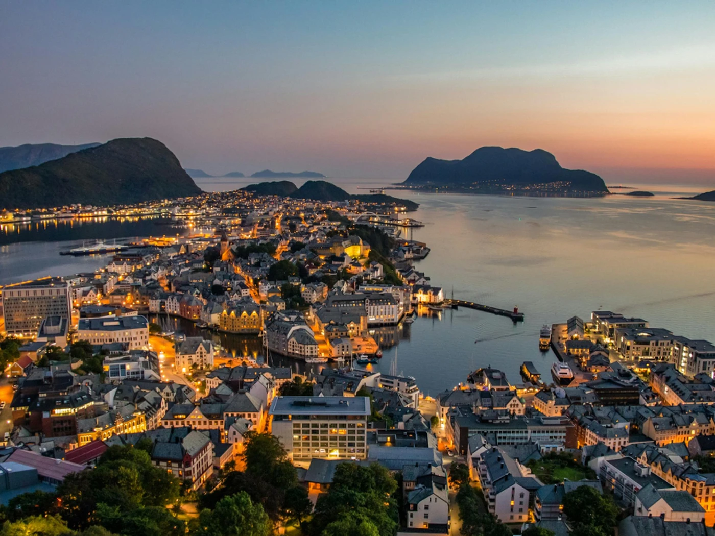Aerial view of a coastal city at dusk, with illuminated buildings, surrounding mountains, and calm waters reflecting the sunset sky.