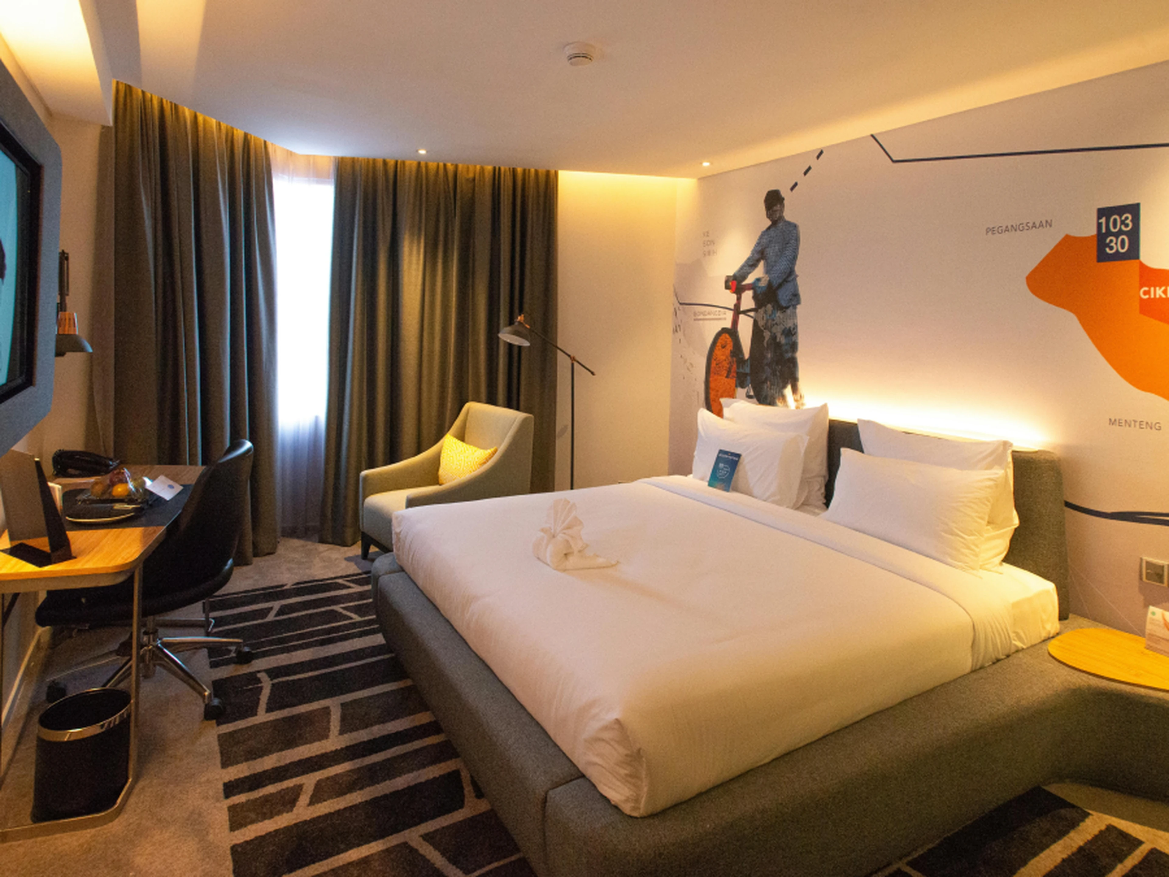 Modern hotel room with a large bed, wall art, desk, chair, and floor lamp. Warm lighting and dark curtains create a cozy atmosphere.