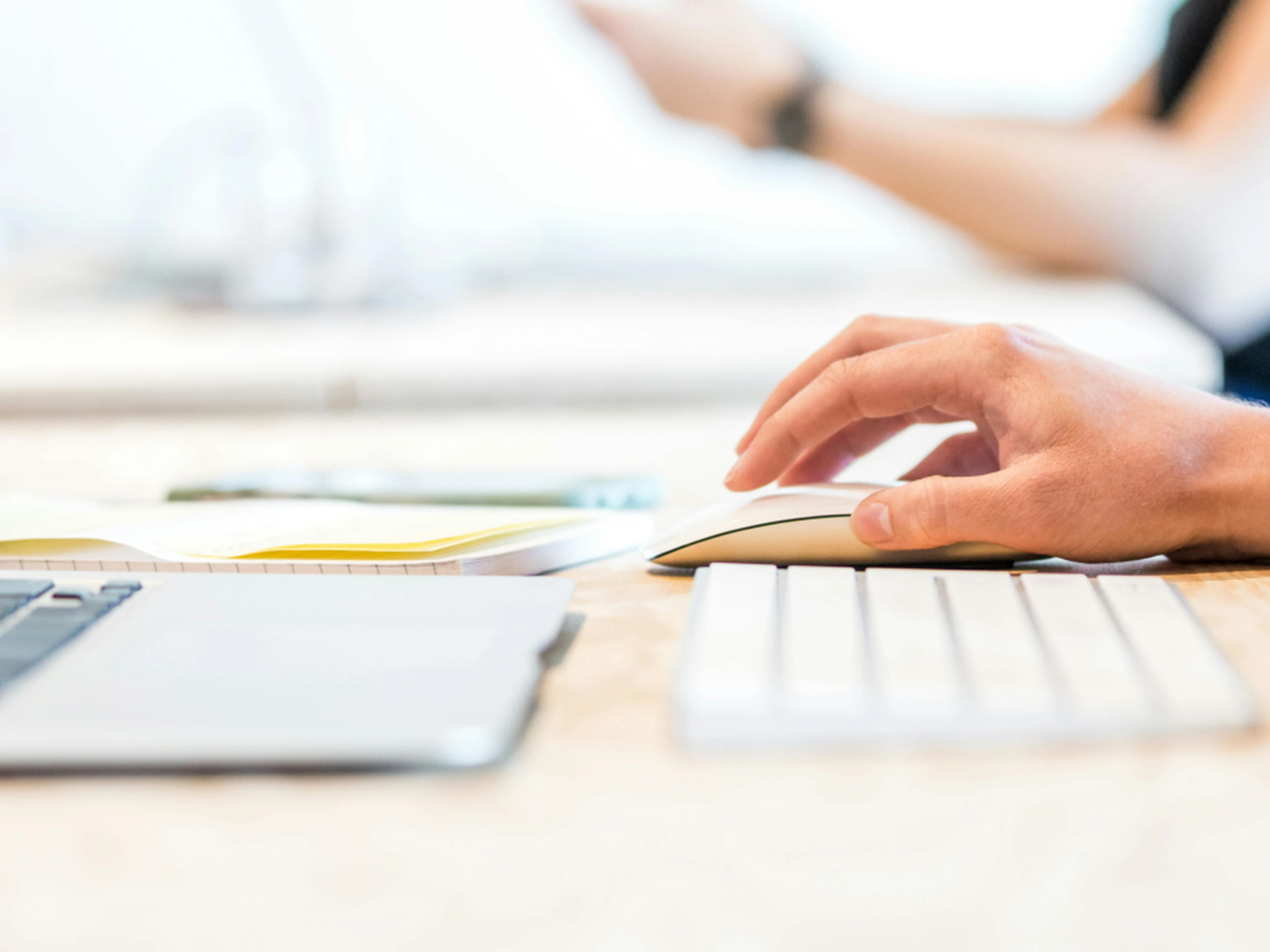 Close-up of a person using a computer mouse on a desk with a keyboard, notebook, and laptop in a bright workspace.