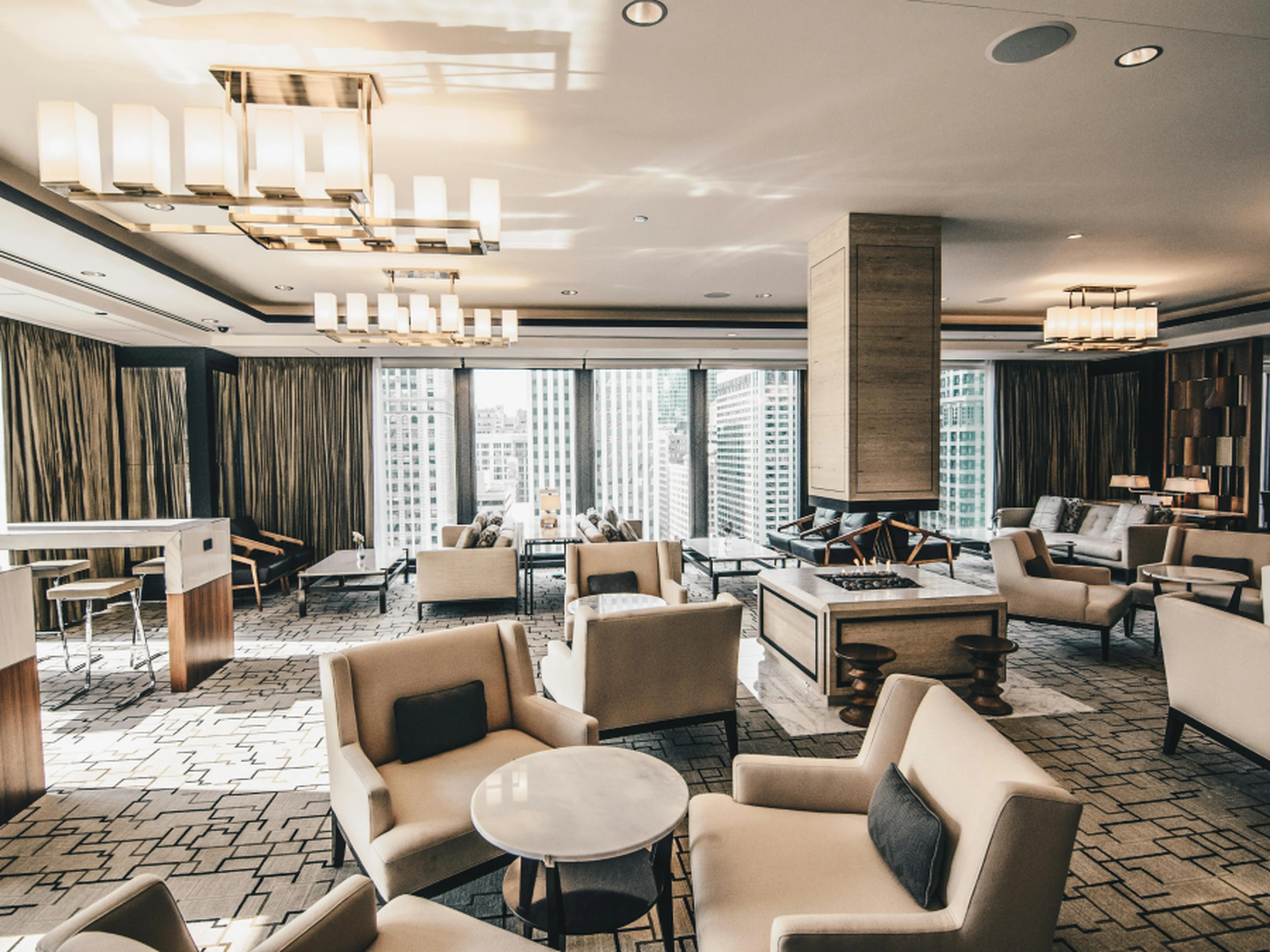 Modern hotel lounge with beige armchairs, round tables, and large windows offering a city view. Contemporary lighting fixtures hang from the ceiling.