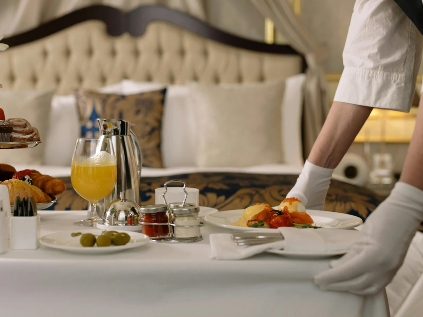 A gloved server arranges breakfast on a hotel room table, featuring eggs Benedict, juice, and pastries, with a luxurious bed in the background.