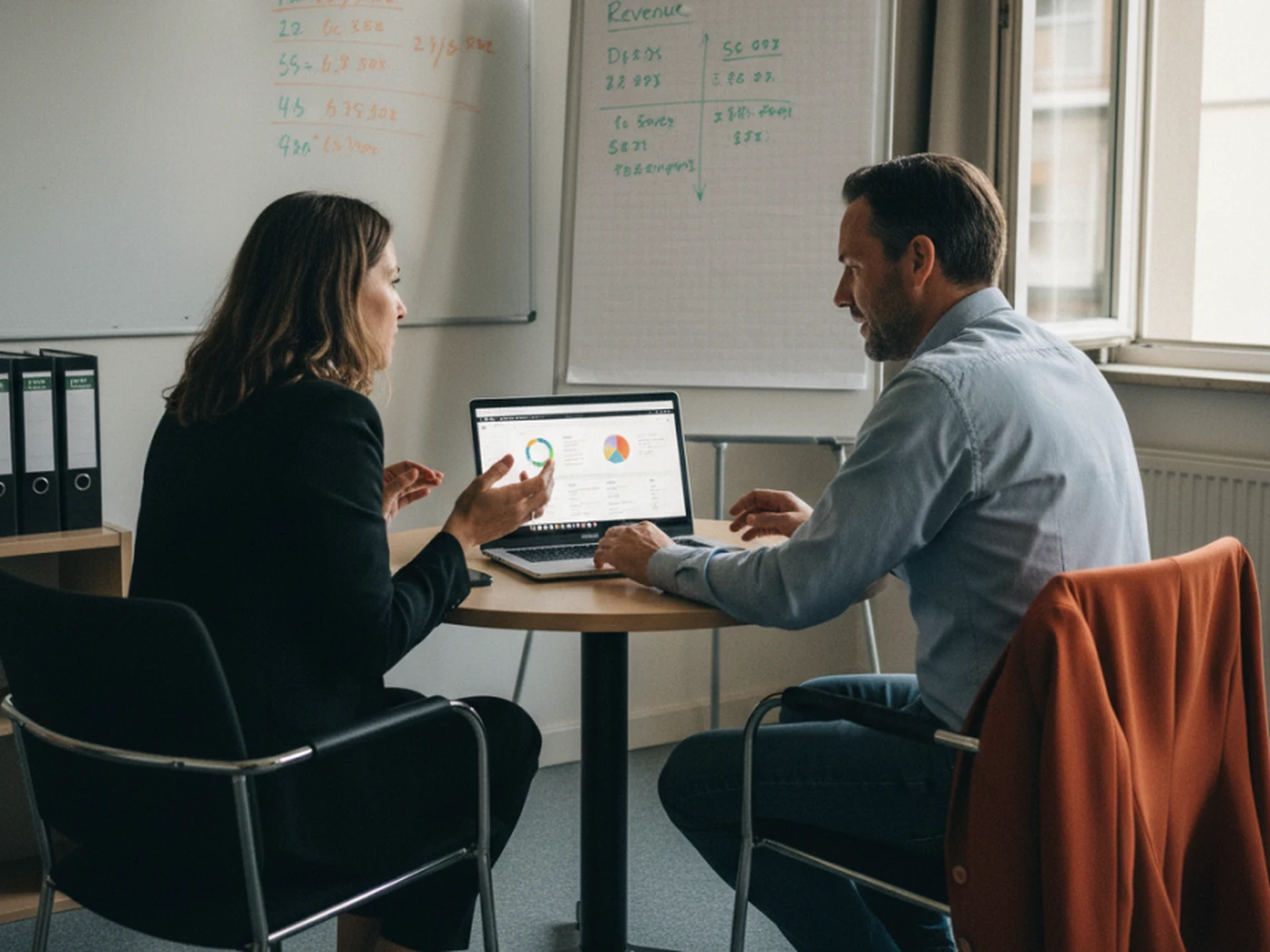 Two people discussing data on a laptop at a small table in an office, with charts on a whiteboard and flip chart in the background.