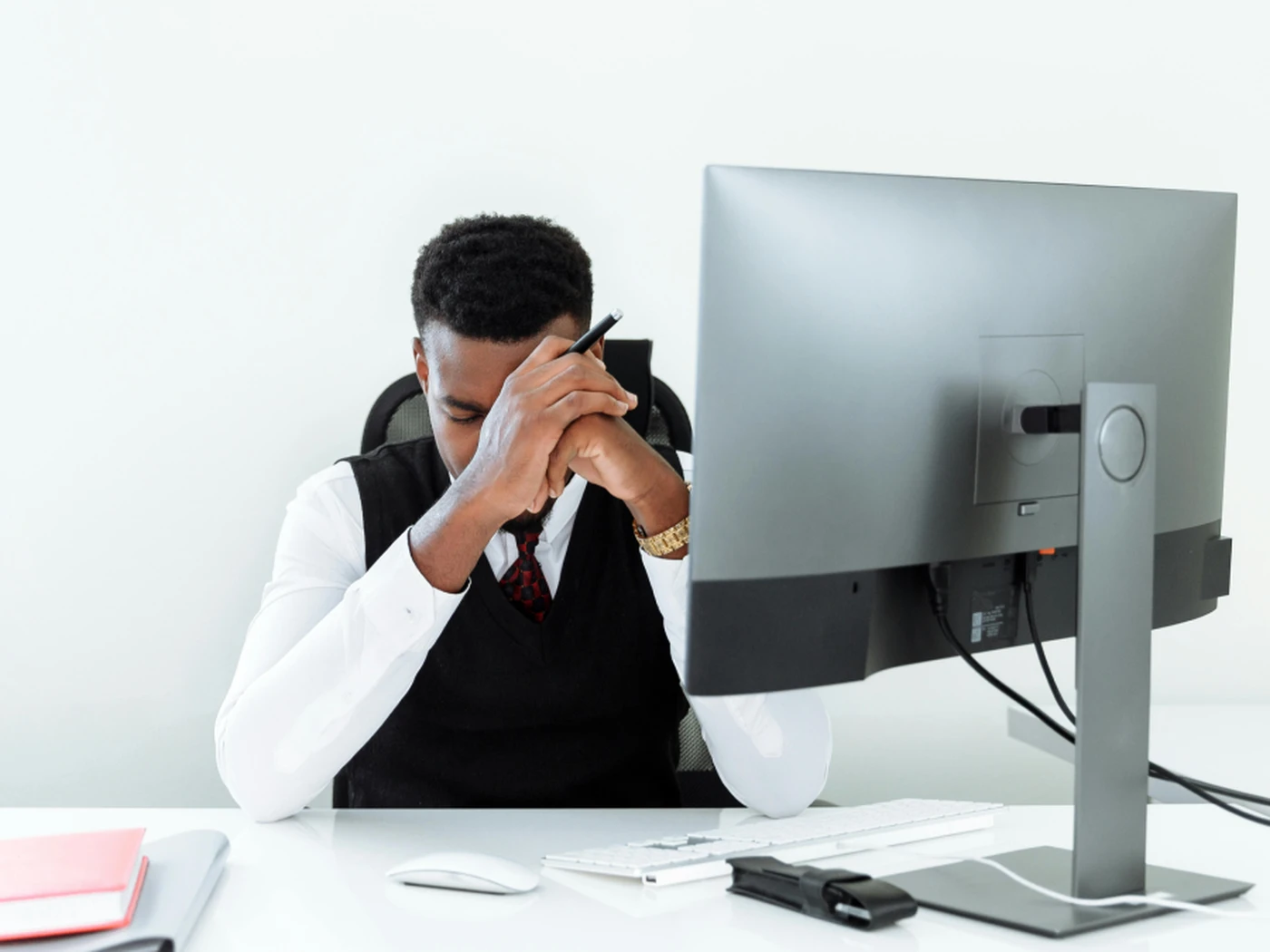 Person sitting at a desk, looking down with hands clasped near their face, in front of a computer monitor in a bright office setting.