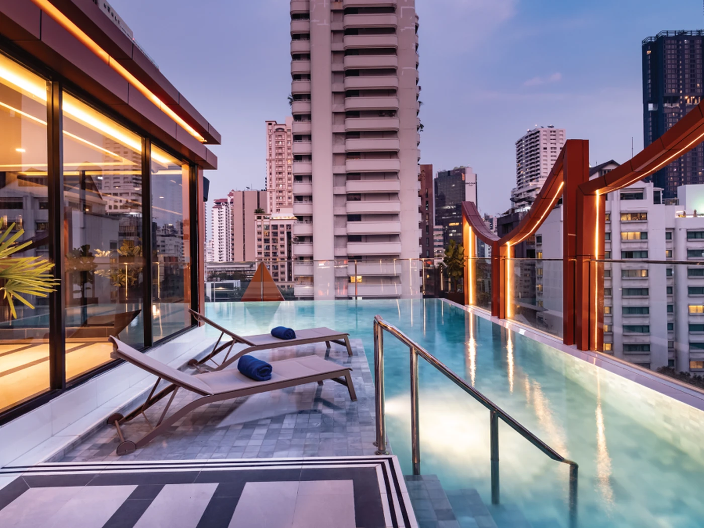 Rooftop infinity pool with two loungers, overlooking a cityscape with tall buildings at dusk.