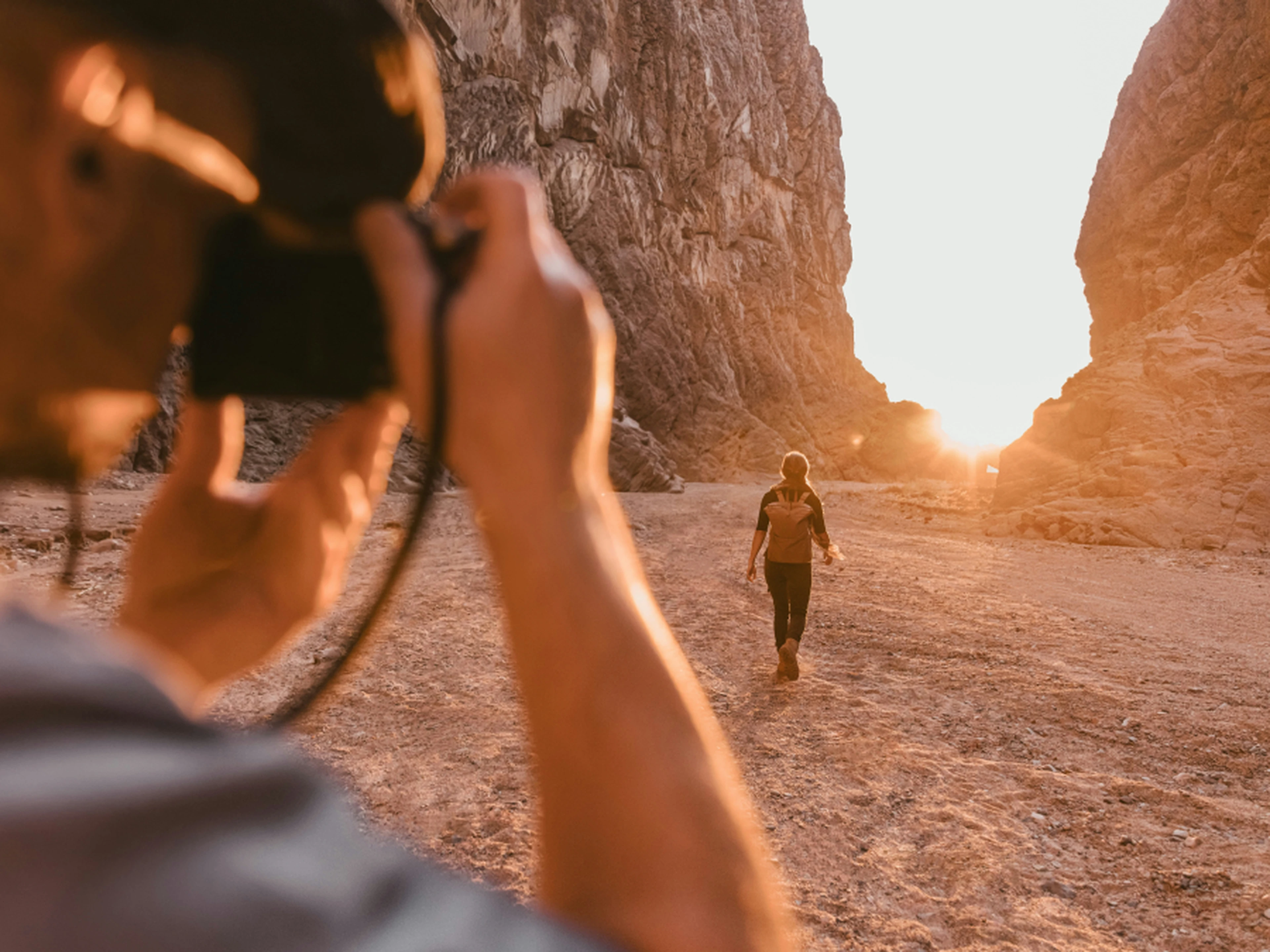 Photographer in foreground photographing a person walking toward the sun through a narrow canyon at sunset