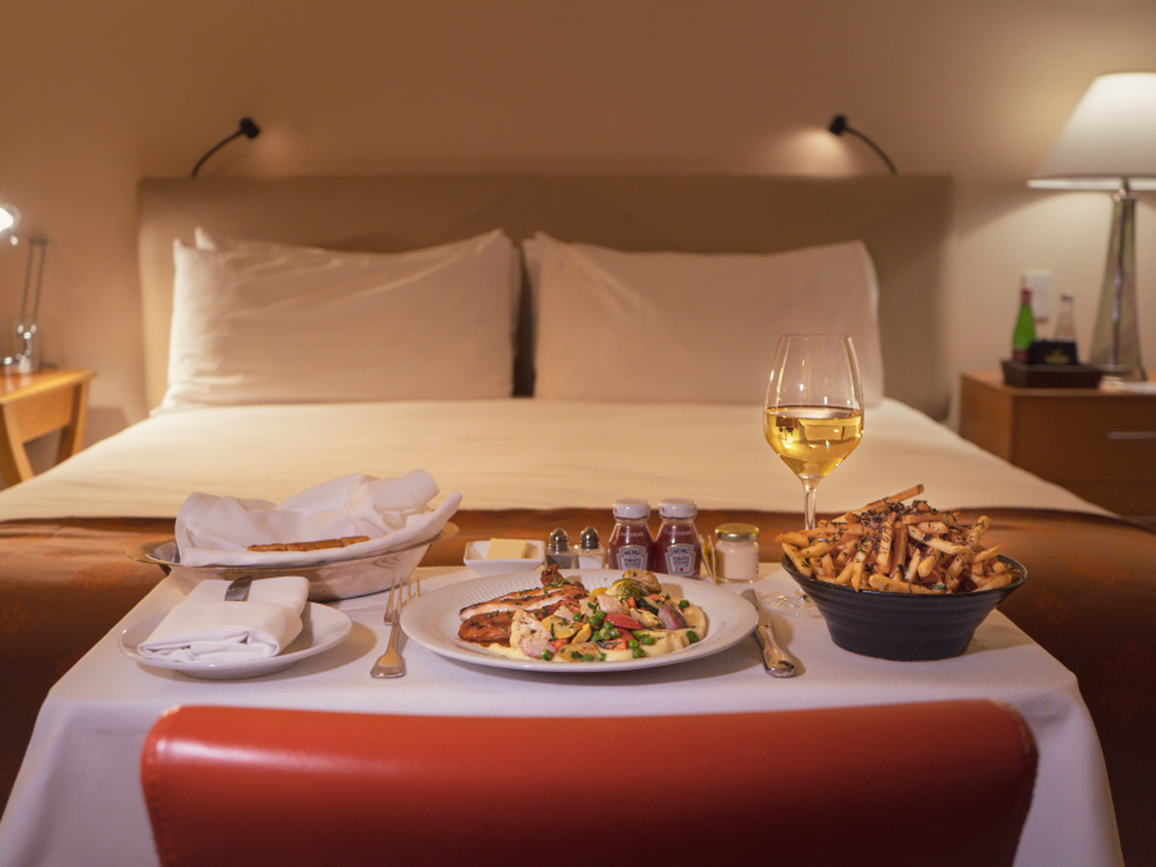 Room service meal with pasta, fries, bread, and a glass of white wine on a table in front of a neatly made bed in a cozy hotel room.