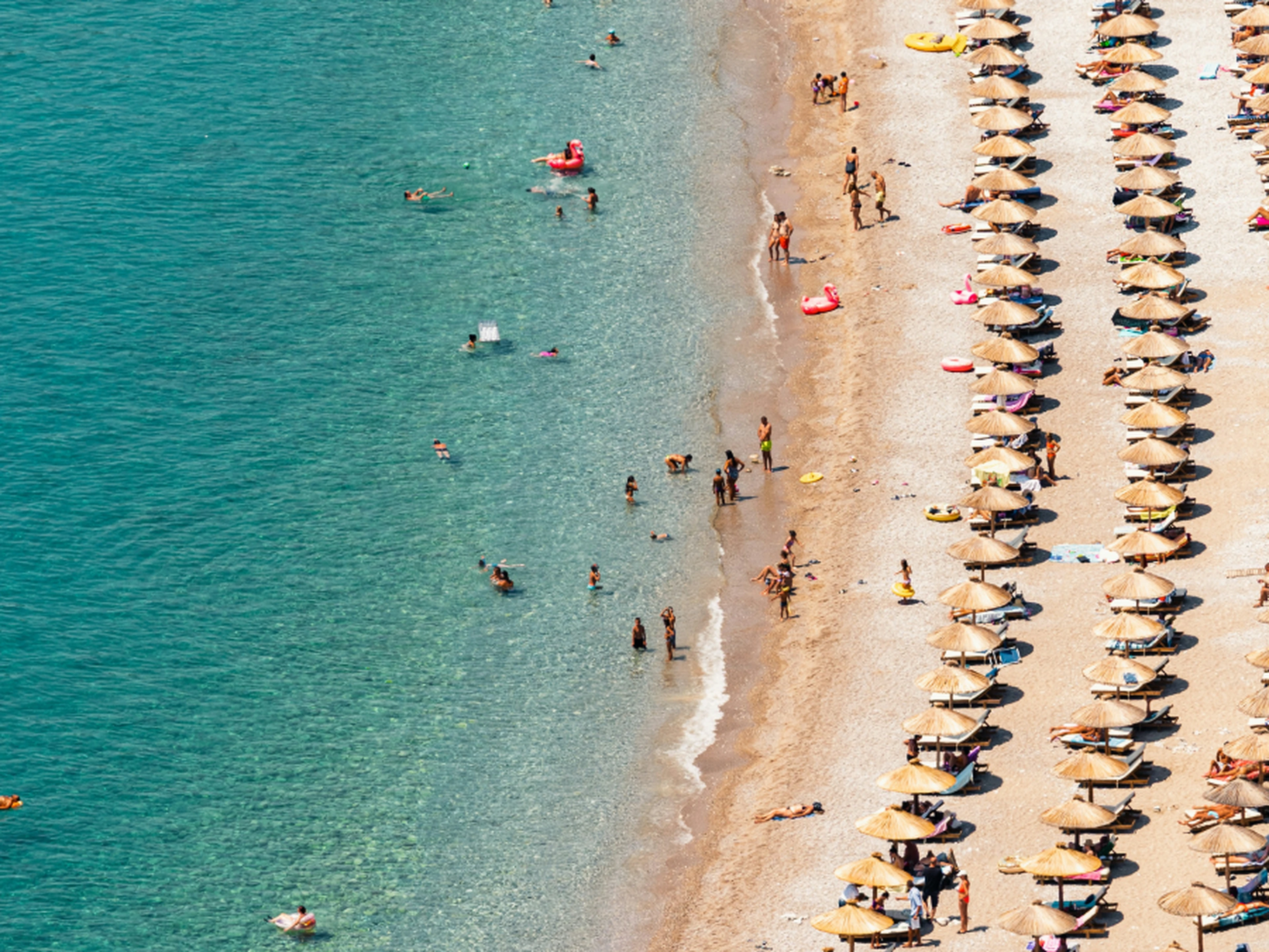 Aerial view of a busy beach with turquoise water, swimmers along the sandy shore and neat parallel rows of straw umbrellas and loungers.