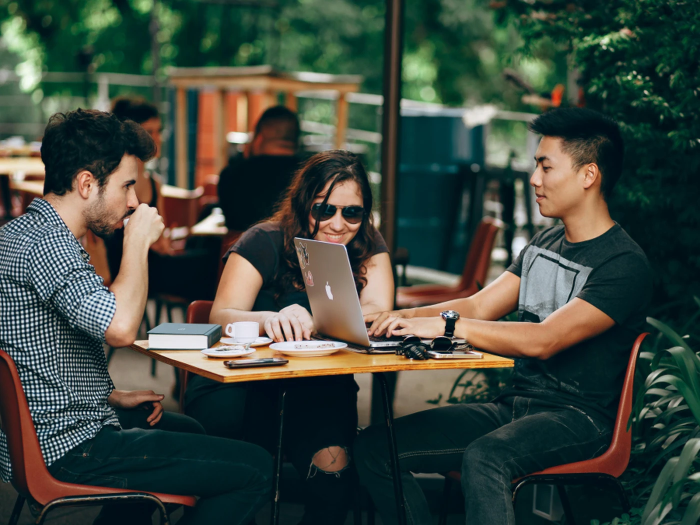Three people sitting at an outdoor café table, engaged in conversation, with a laptop and coffee cups on the table.