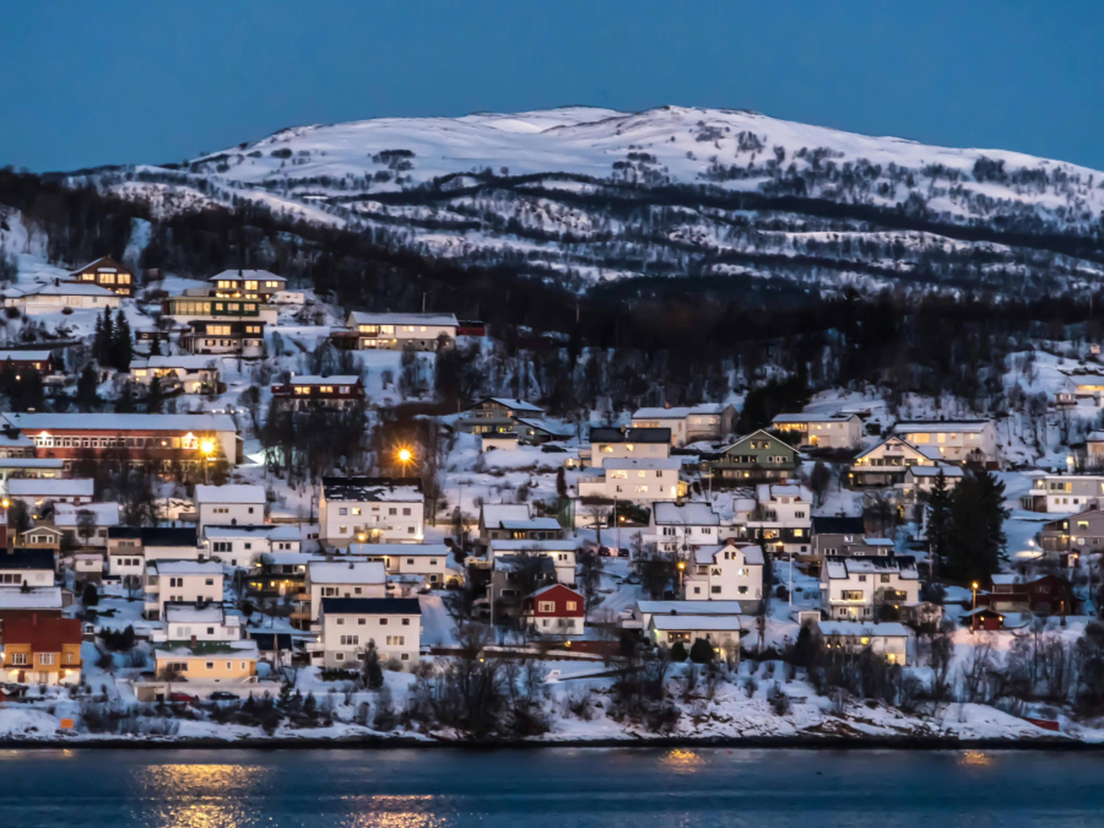 Snow-covered houses on a hillside at dusk, with glowing windows and a snowy mountain in the background, reflected in a calm body of water.