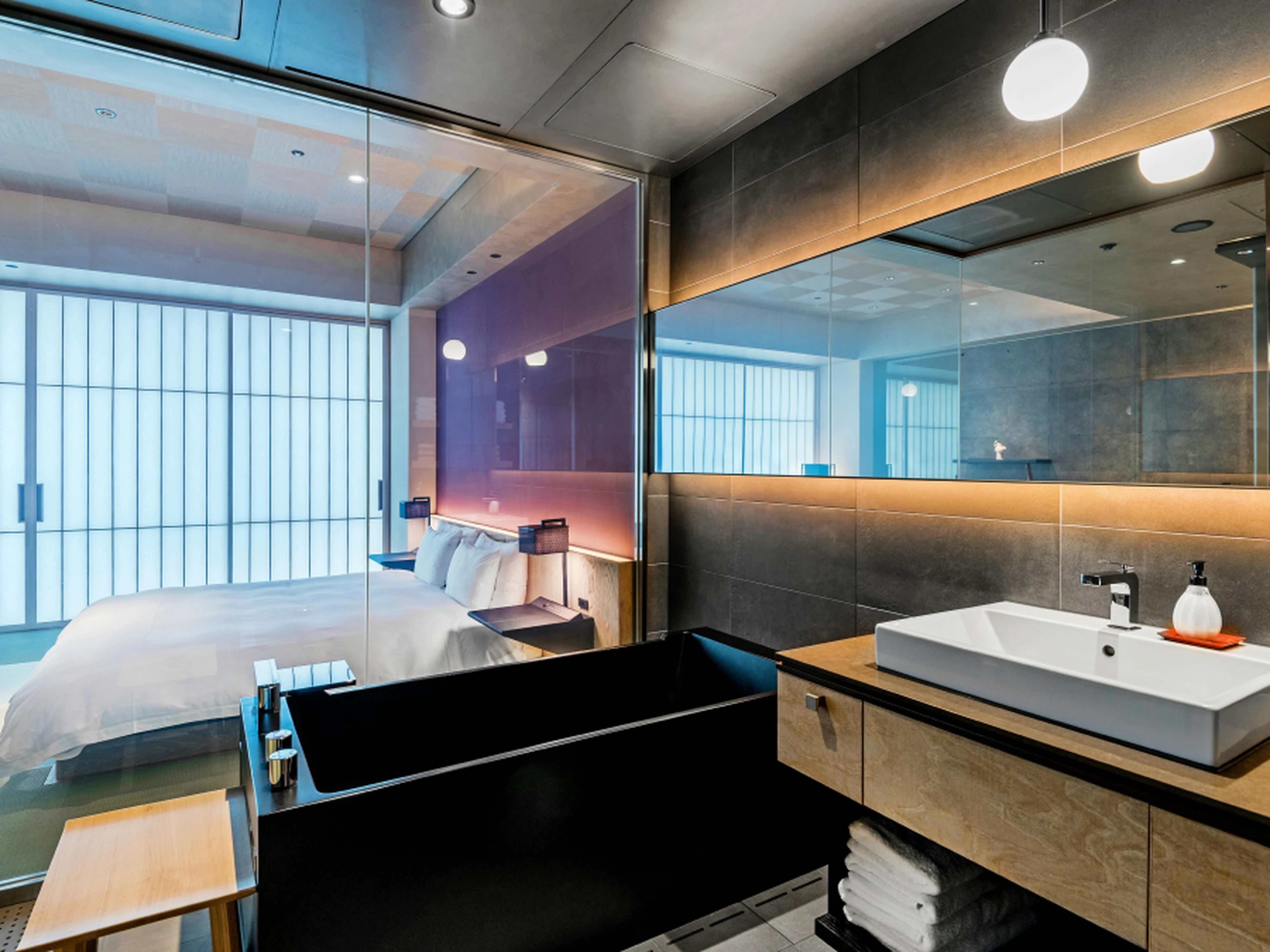 Modern hotel room with a glass-walled bathroom, featuring a sleek black bathtub, white sink, and view of a cozy bed with soft lighting.