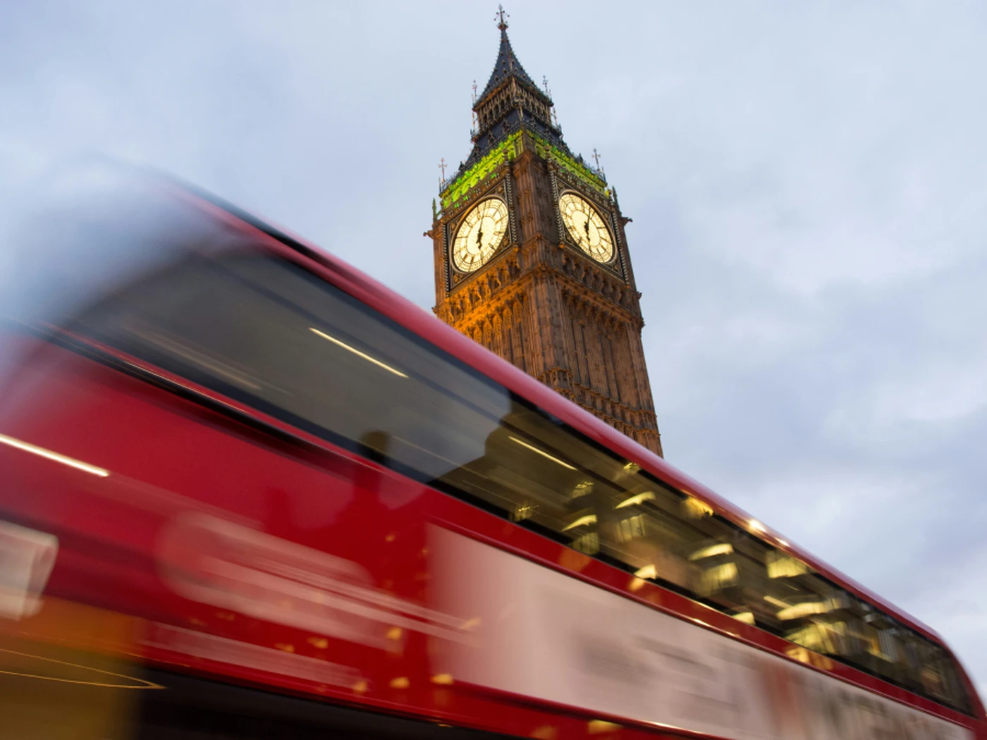 A blurred red double-decker bus passes in front of the illuminated Big Ben clock tower in London, set against a cloudy sky.
