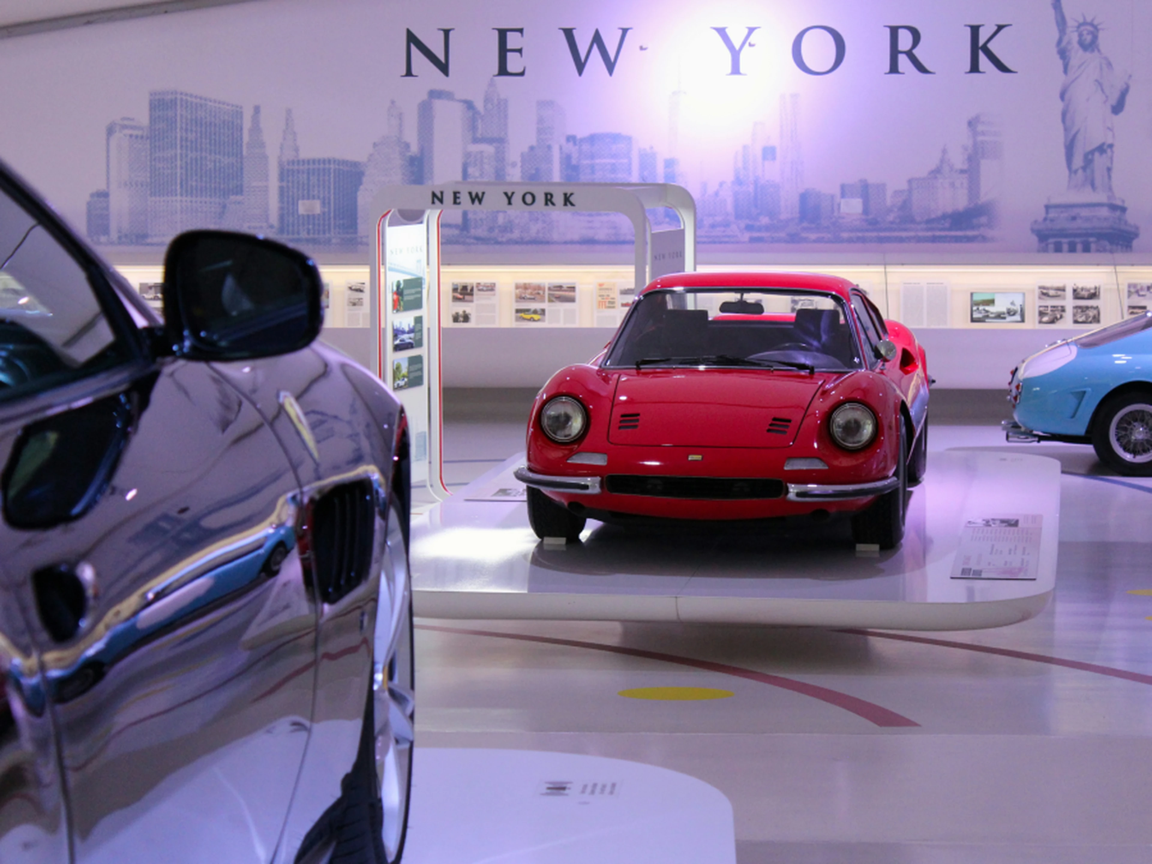 A red vintage car displayed in a showroom with a New York theme, featuring cityscape images and other cars in the background.